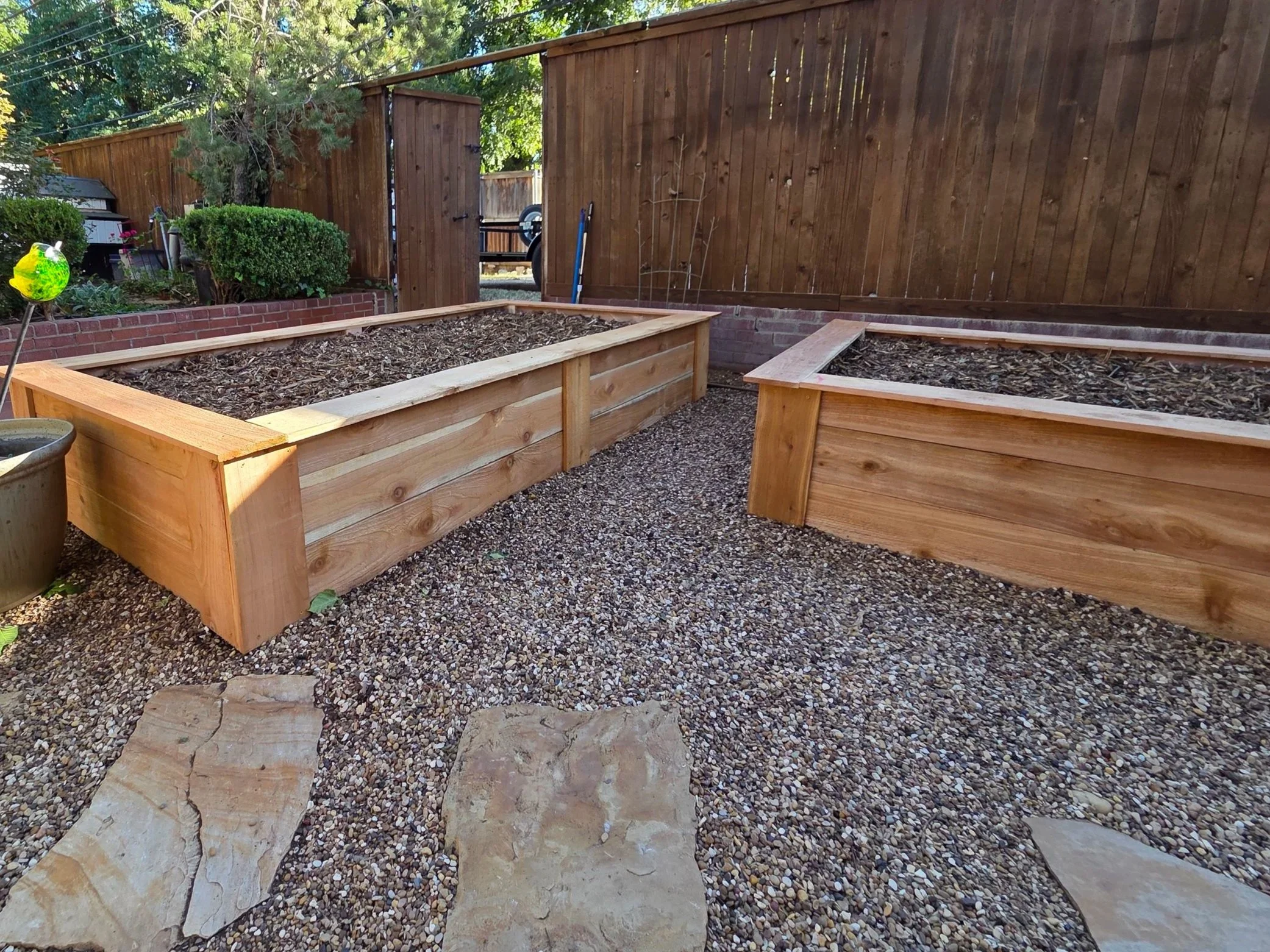 Two wooden raised garden beds filled with soil, situated in a backyard with a gravel ground, wooden fence, and trees in the background.