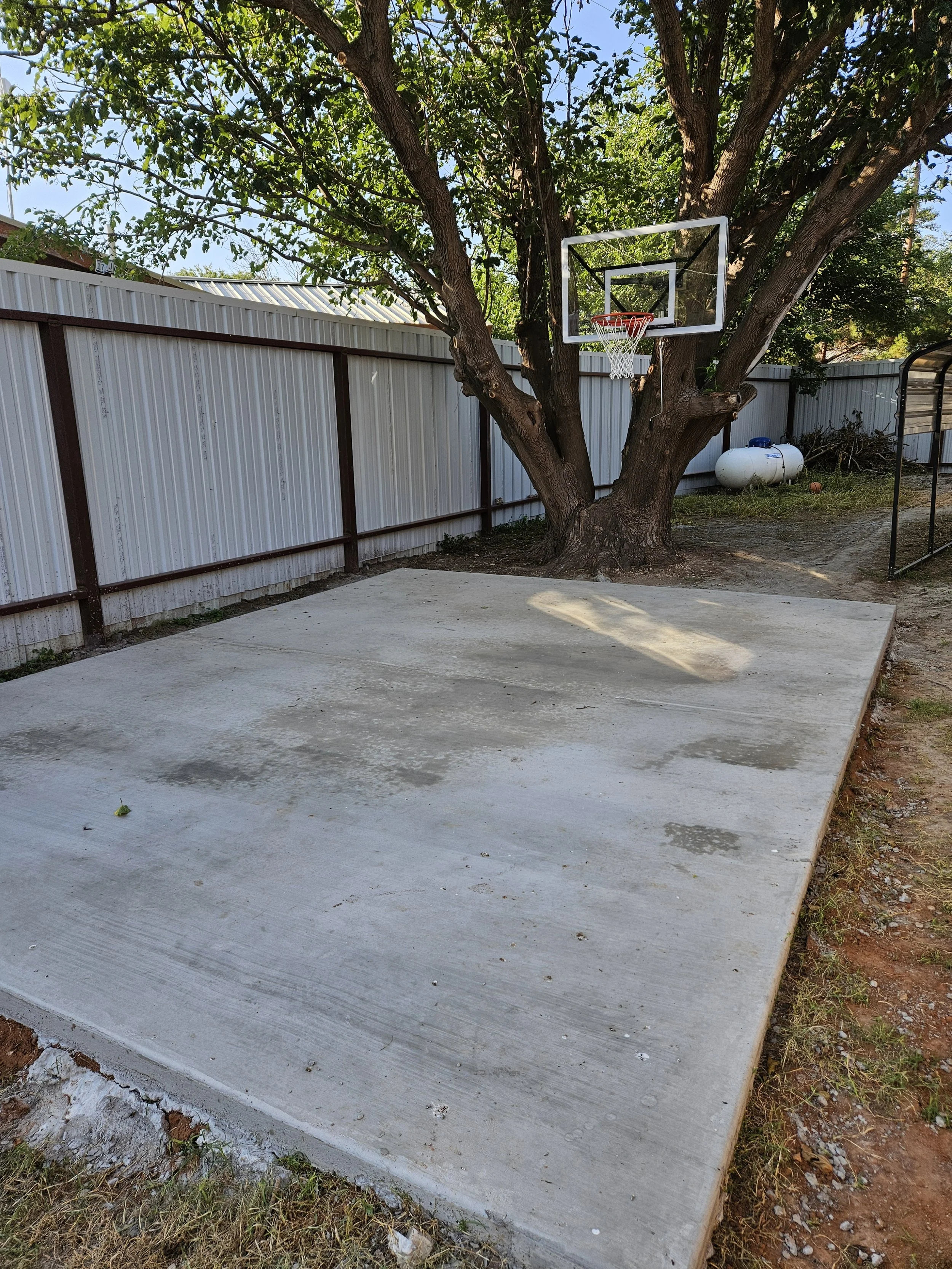 Concrete basketball court with a basketball hoop mounted on a tree, with a white metal fence and some yard items in the background.