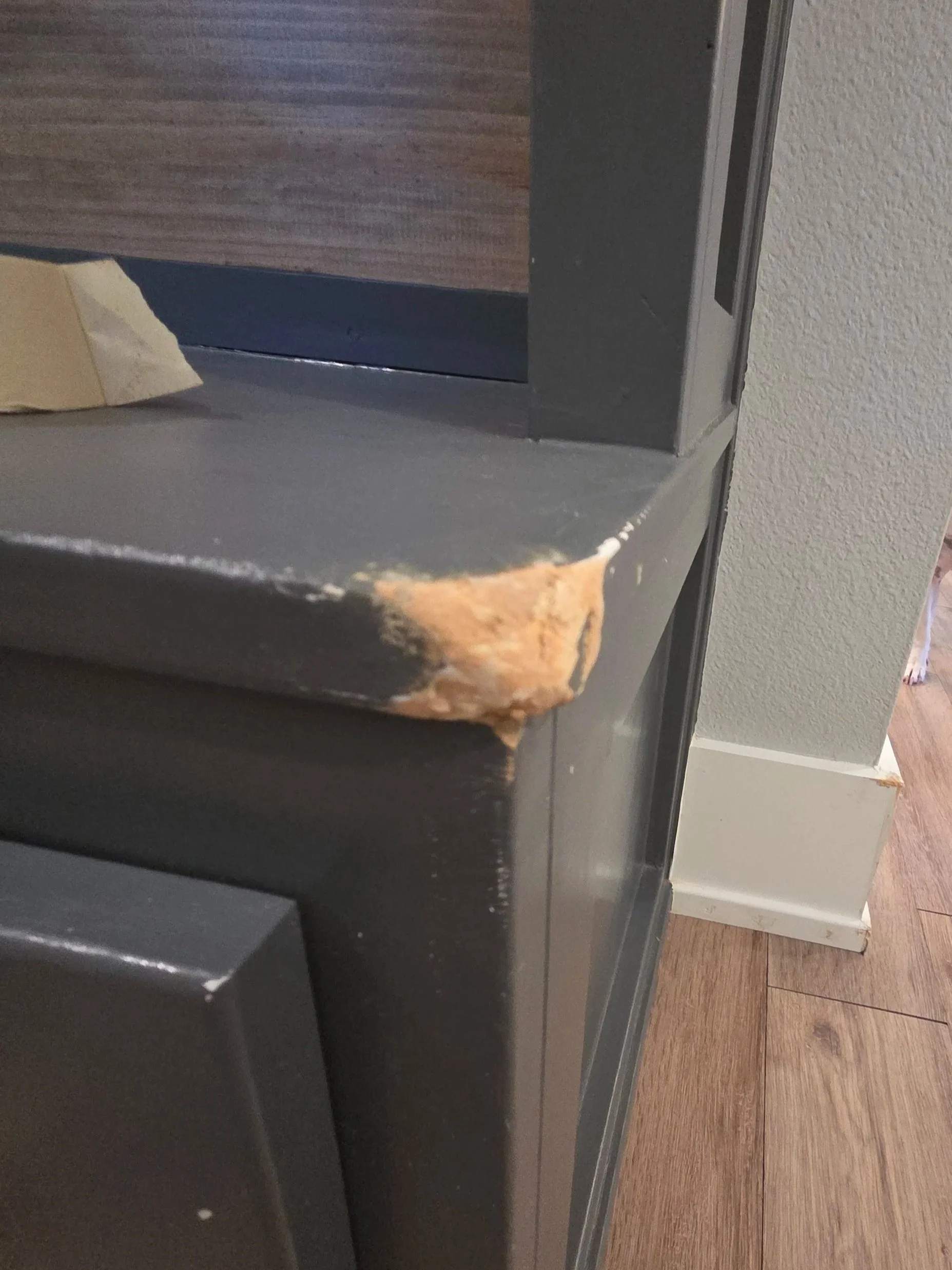 Close-up of a black piece of furniture with a chipped corner revealing damaged wood underneath, next to a gray wall and wooden flooring.