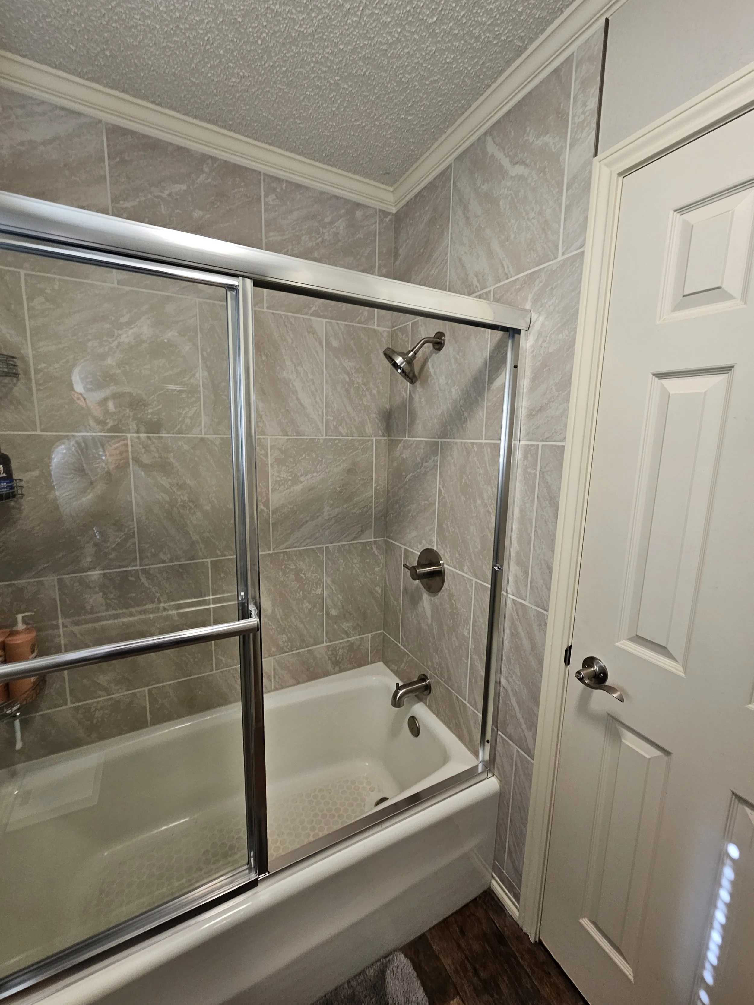 Bathroom shower with beige stone tiles, two metal showerheads, and a sliding glass door. There is a white bathtub and partially visible white closet door.