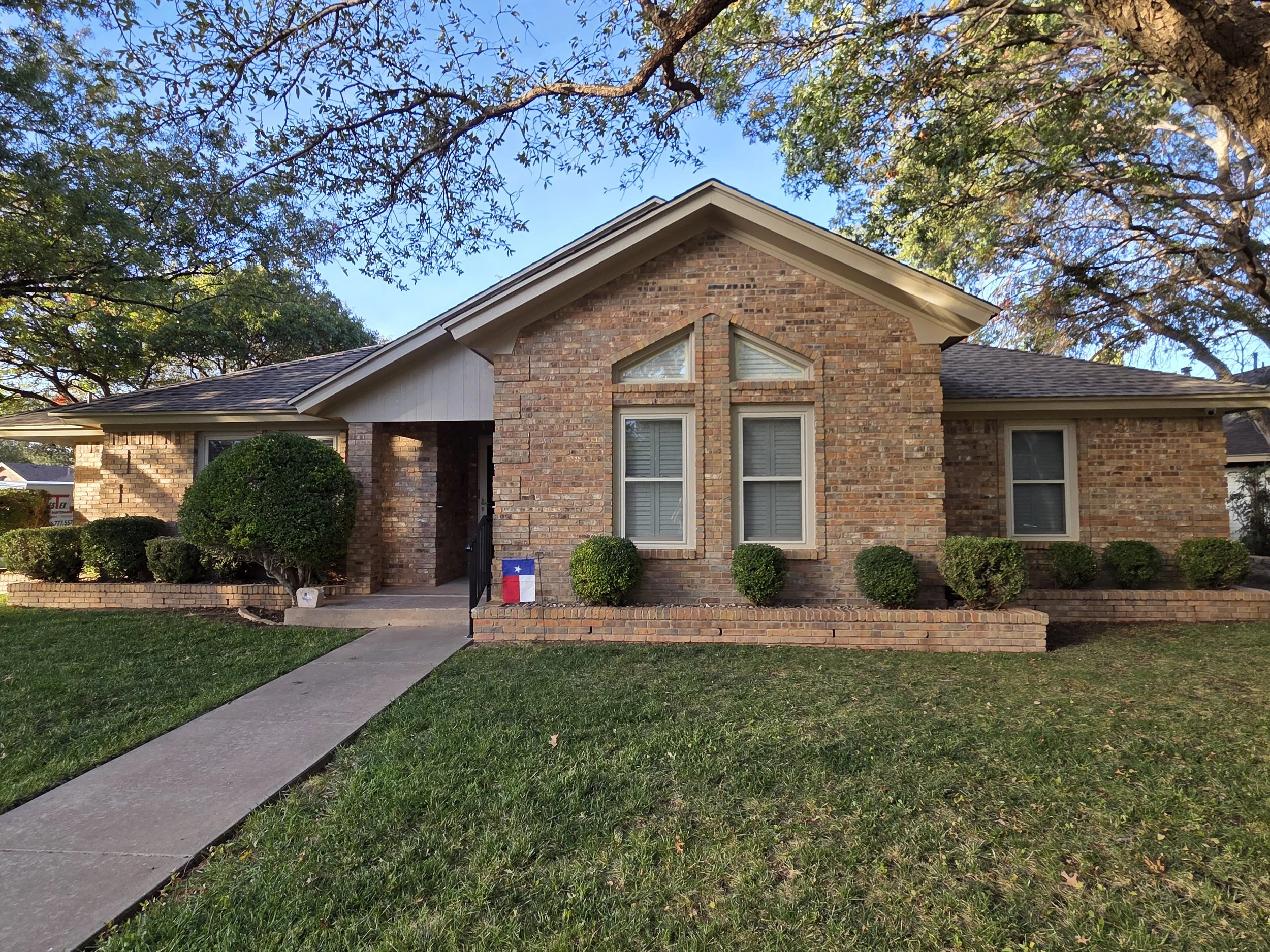 A brick house with a gabled roof and three windows in the front, surrounded by manicured bushes and a green lawn, with trees overhead and a sidewalk leading to the front entrance.
