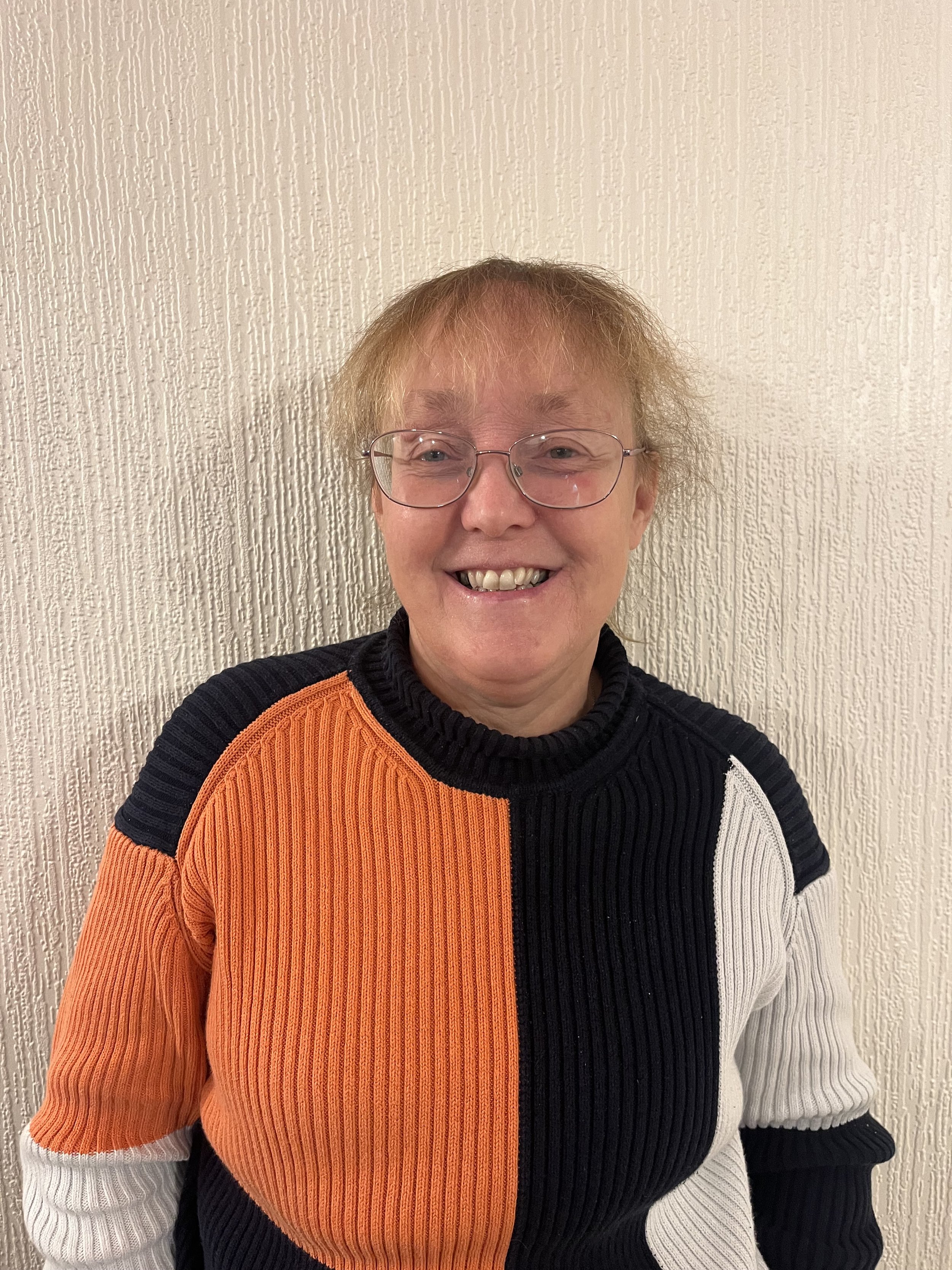 A smiling woman with glasses and short, light-colored hair standing in front of a textured beige wall, wearing a color-blocked sweater with orange, black, white, and navy sections.
