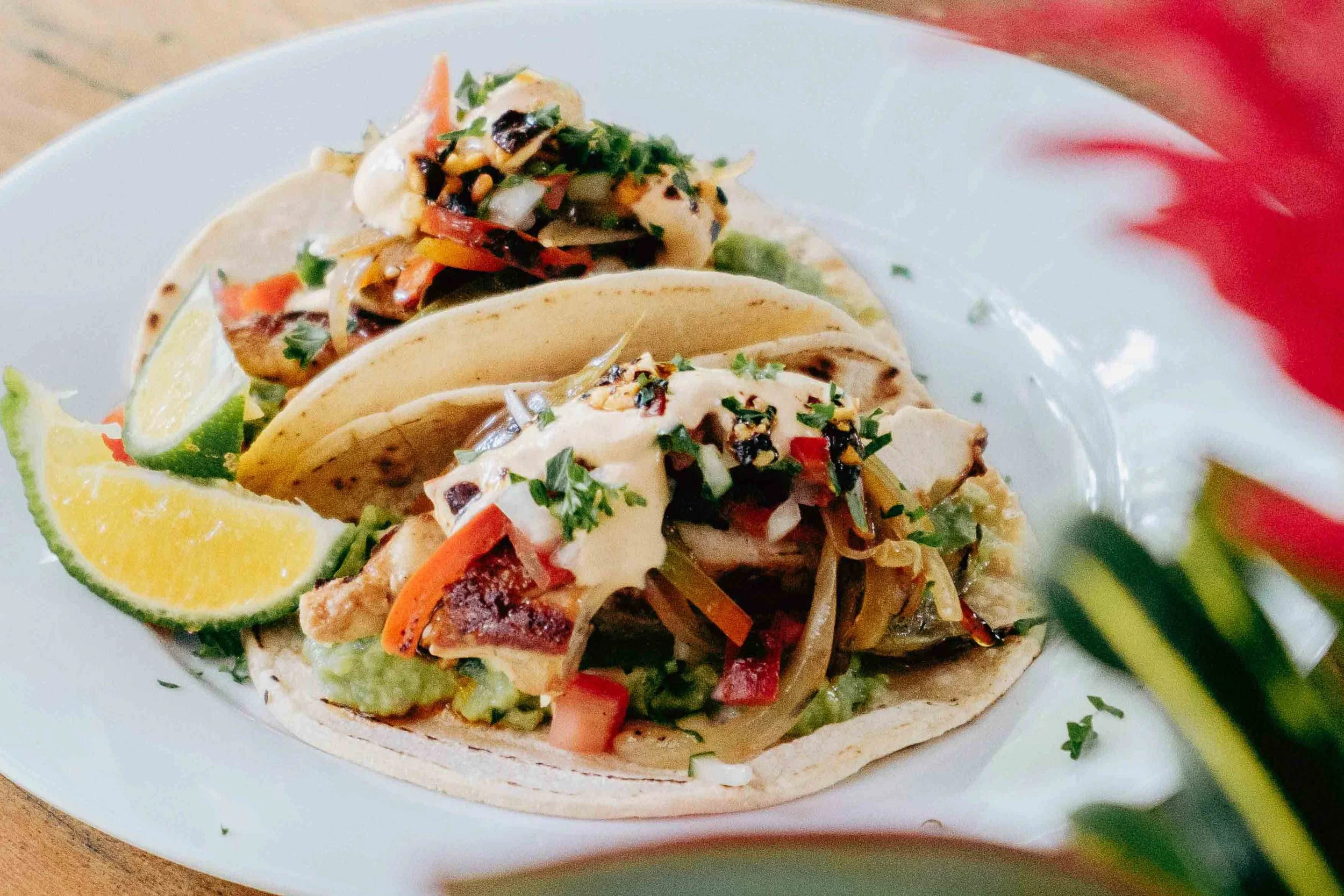 Two tacos on a white plate garnished with lime wedges, filled with vegetables, meat, and topped with sauce and herbs.
