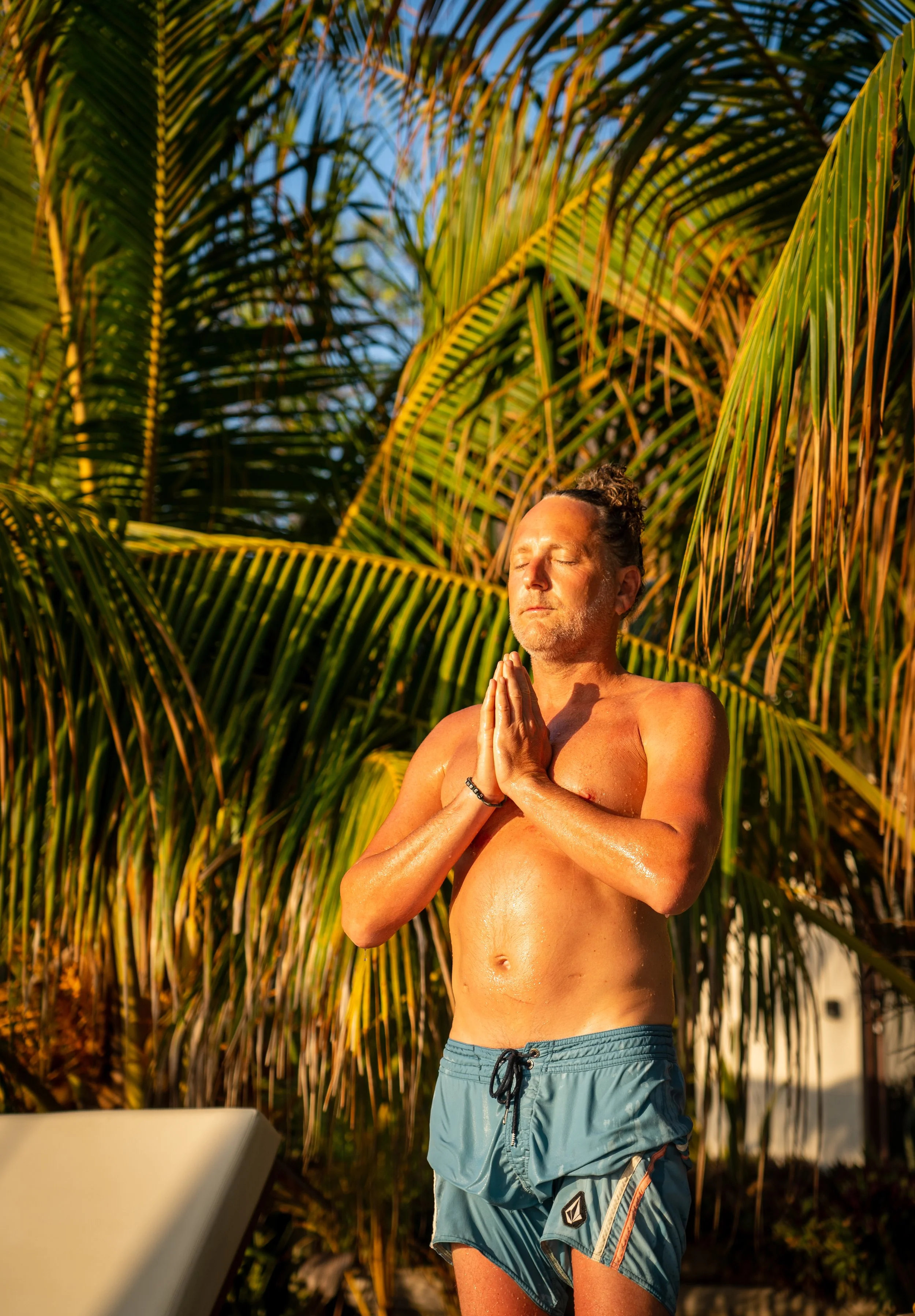 A man practicing yoga outdoors among palm trees, with his hands in prayer position, eyes closed, basking in sunlight.