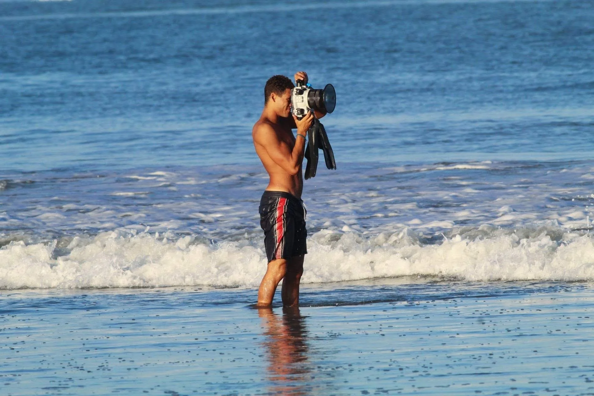 A shirtless man standing in the ocean, holding a camera and taking photos, with waves crashing around him.
