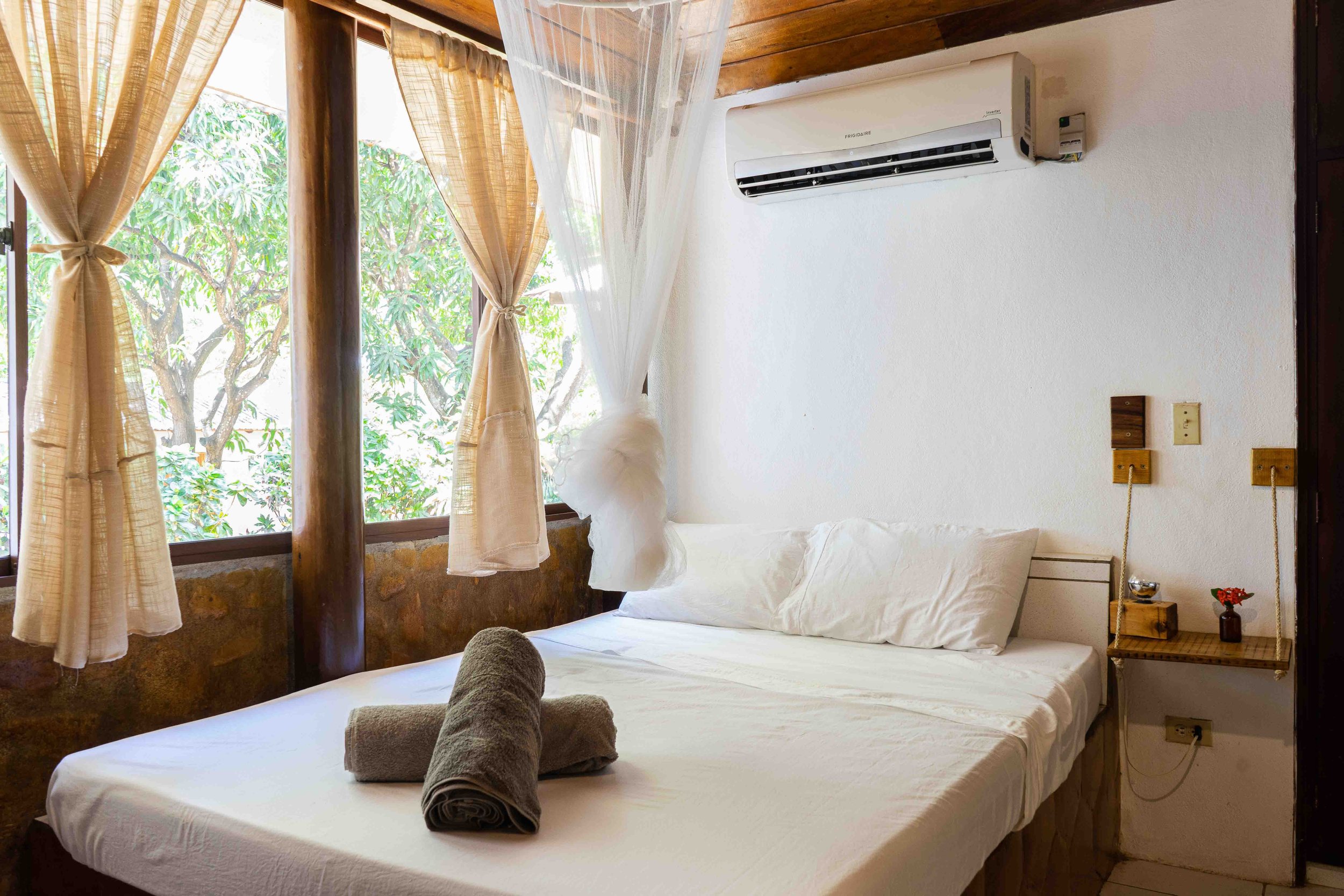 Cozy bedroom with a large bed, rolled towels, a shelf with a small flower, and a window with sheer curtains overlooking a garden.