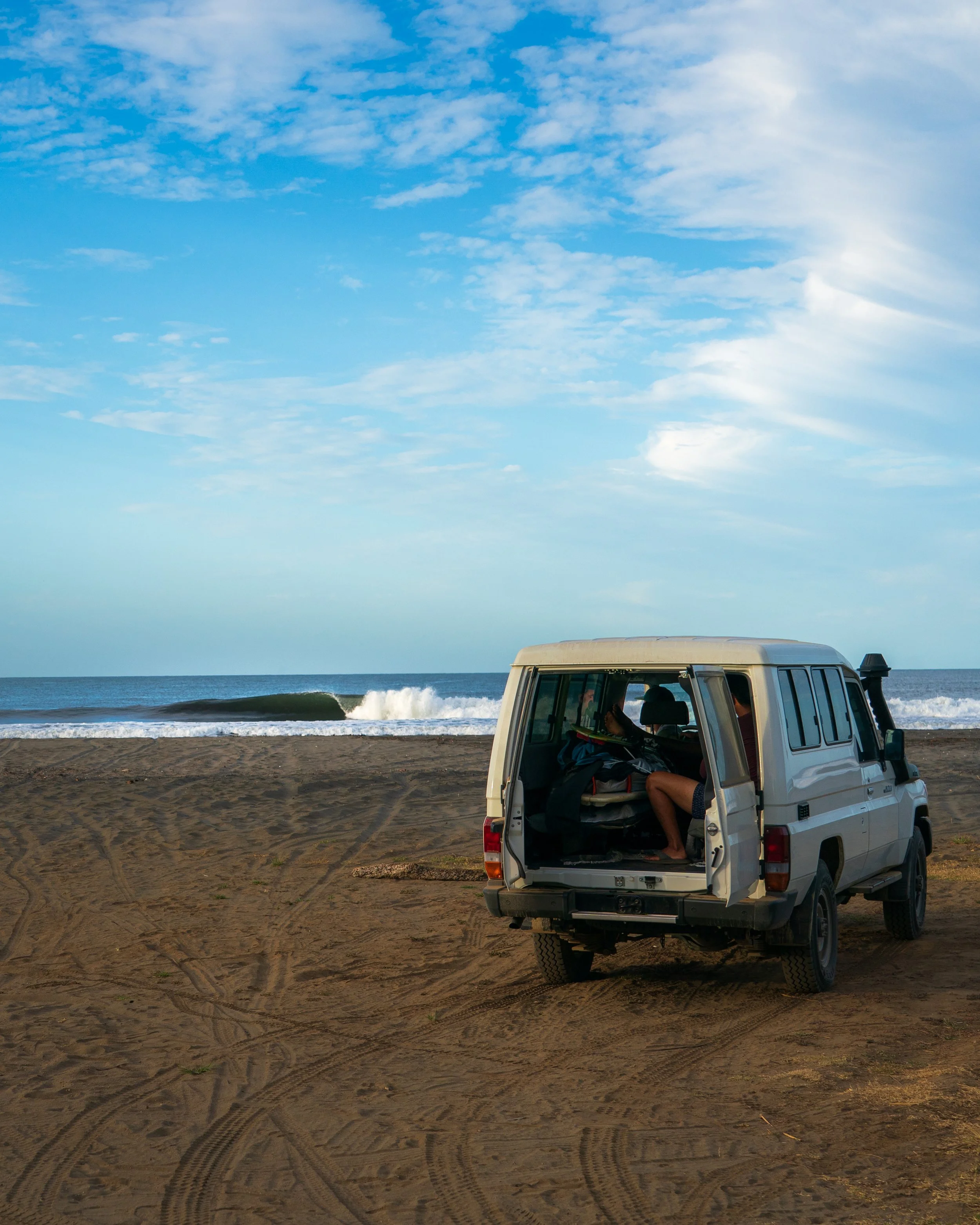 A white vehicle parked on a sandy beach with the ocean and waves in the background and a partly cloudy sky above.