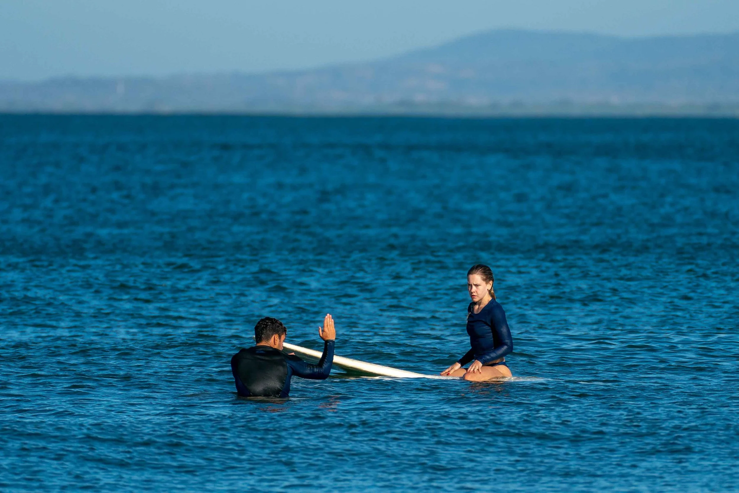 A man and woman in wetsuits in the ocean with a surfboard, woman sitting on her knees and man giving surf coaching tips