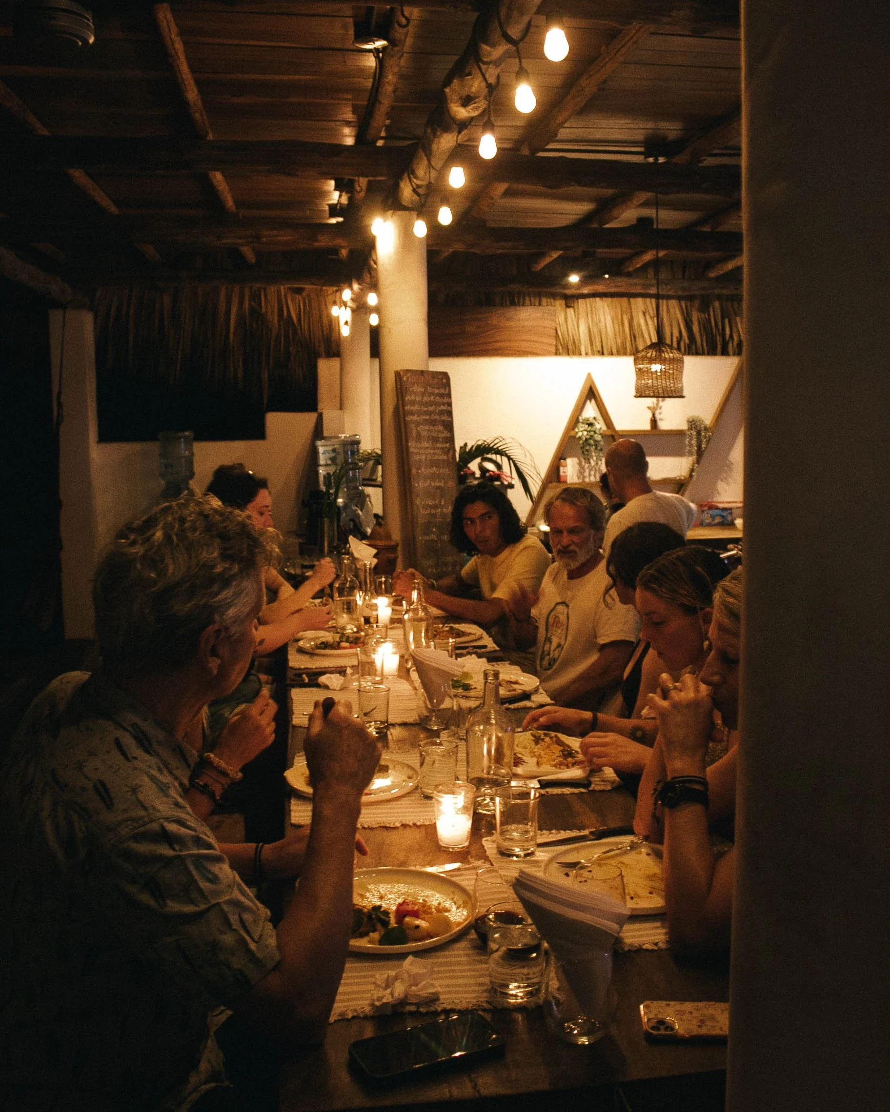People gathered around a long dining table in a dimly lit restaurant, enjoying a meal with candles on the table, rustic decor with warm lighting, and a chalkboard menu in the background.