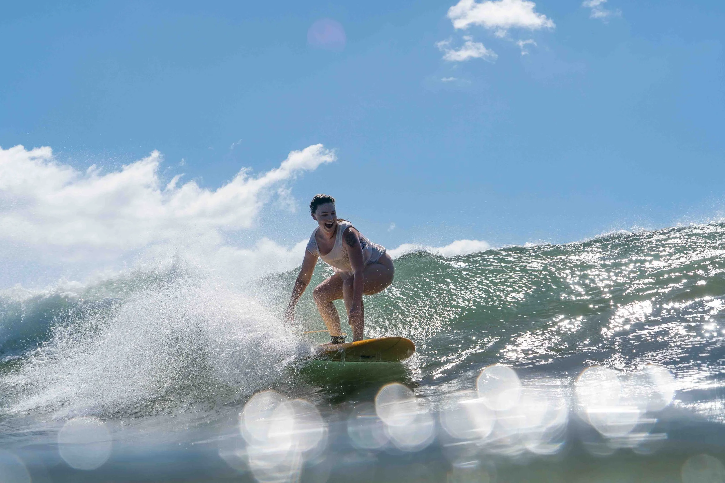 Woman surfing on a wave with a smile on her face under a blue sky with scattered clouds.