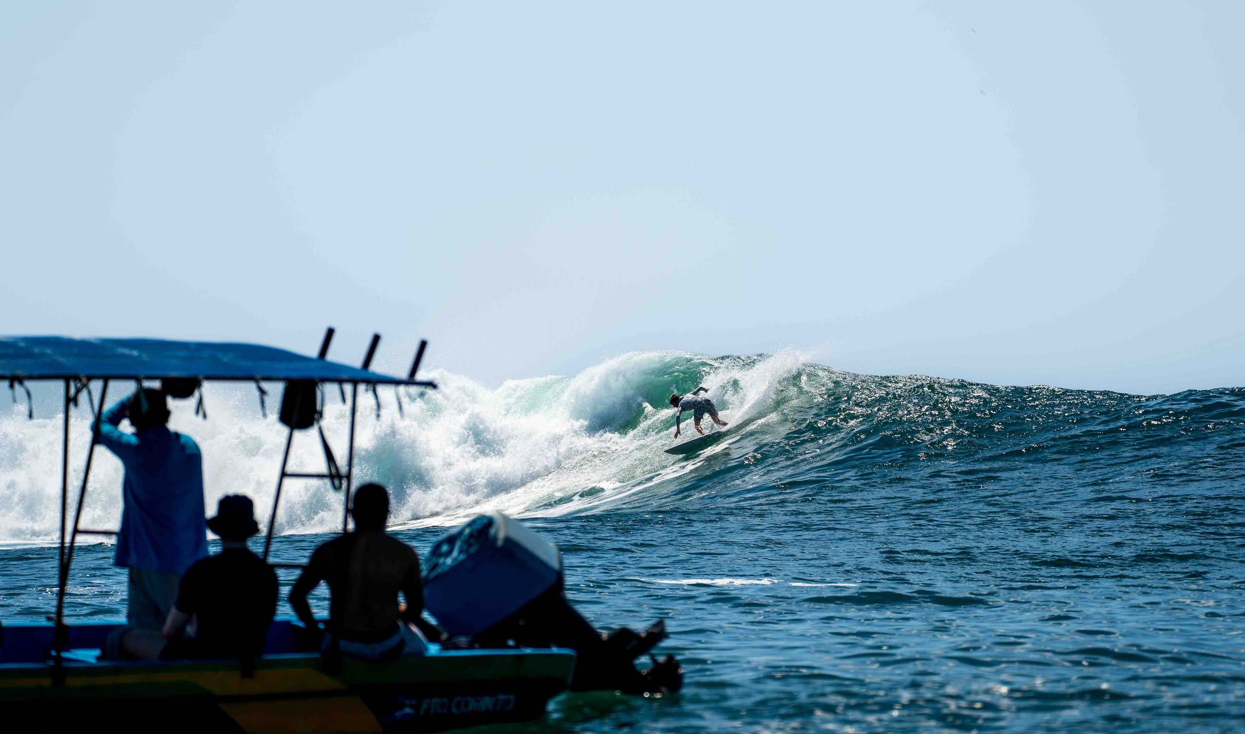 A surfer riding a wave in the ocean with three people on a boat watching in the foreground.