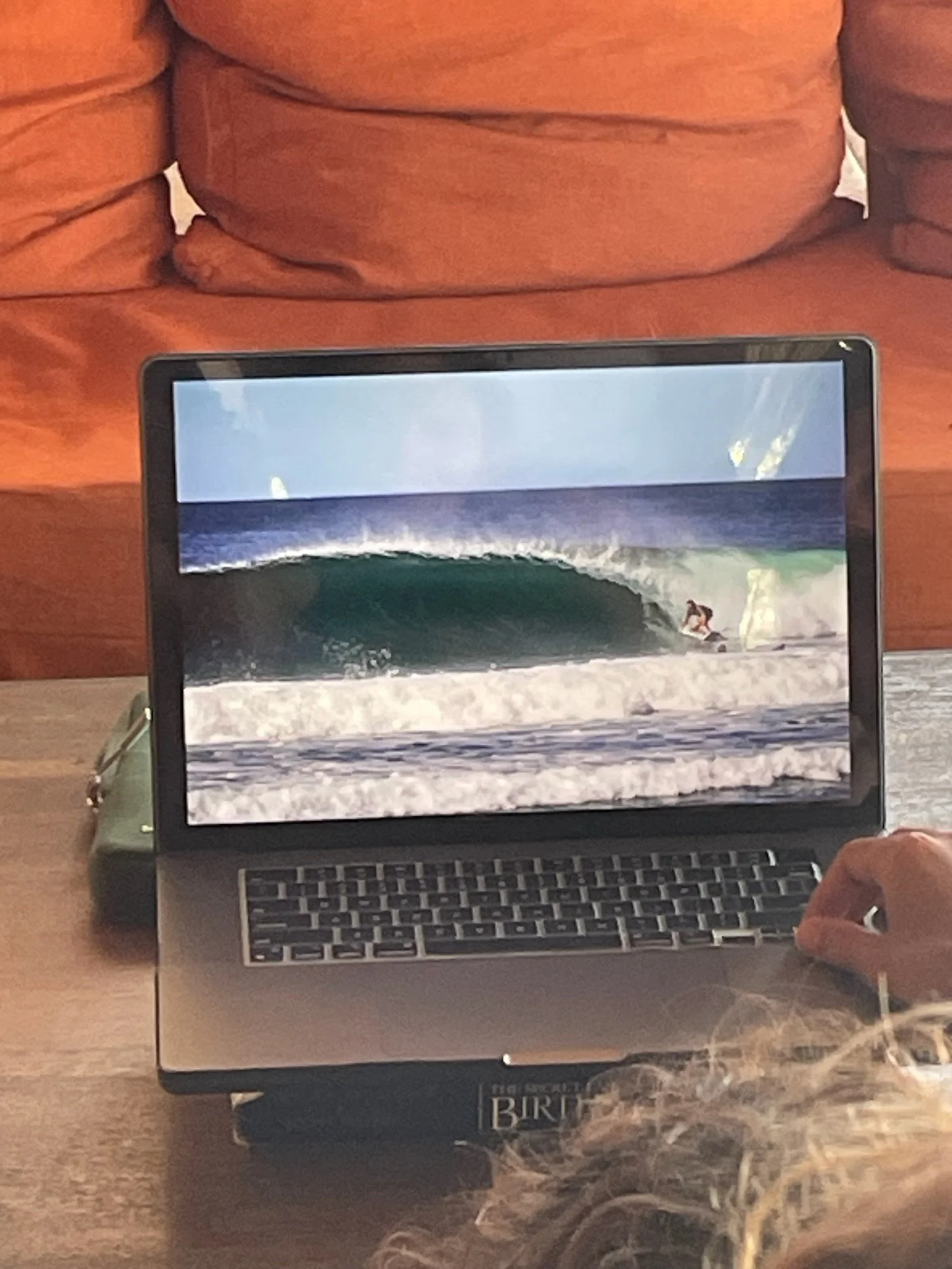 Laptop on a desk showing a surfer riding a wave at the beach with a person in curly hair in the foreground.