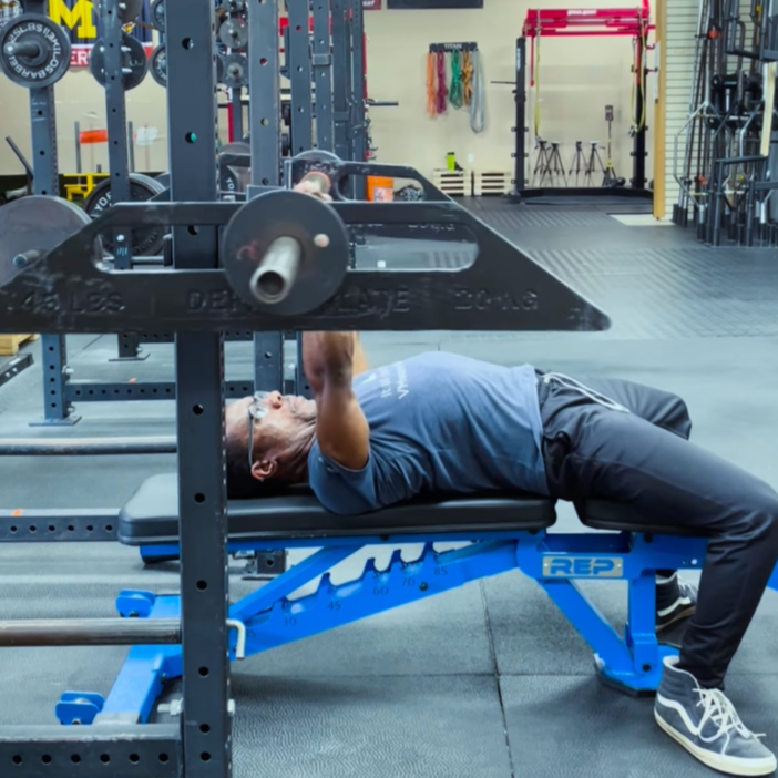 An elderly black man lifting weights on a bench press in a gym. The weights are a innovative shape and rock creating instability.