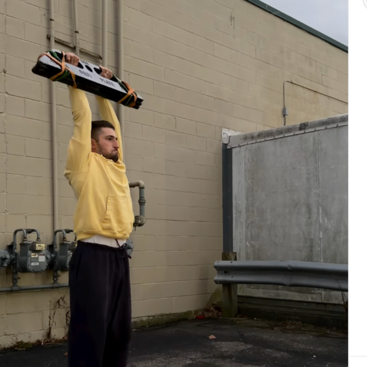 Man in yellow hoodie standing outdoors next to a beige brick wall, doing a strongman block press above his head with both hands.