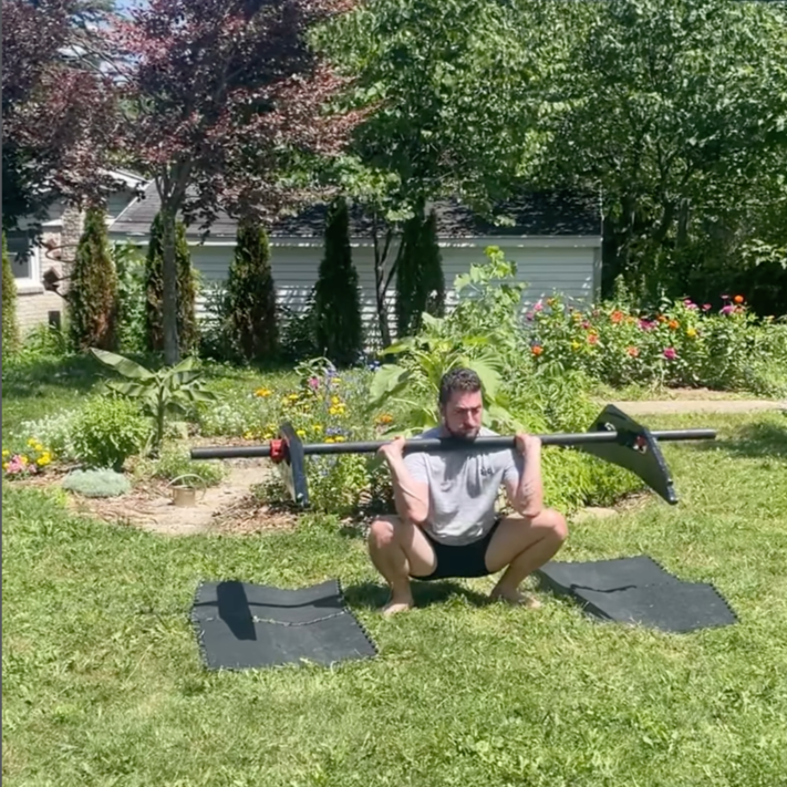 A man exercises outdoors in a park or garden, doing a squat with a long barbell resting on his shoulders, with mats on the grass beneath him and a colorful flower garden and trees in the background.