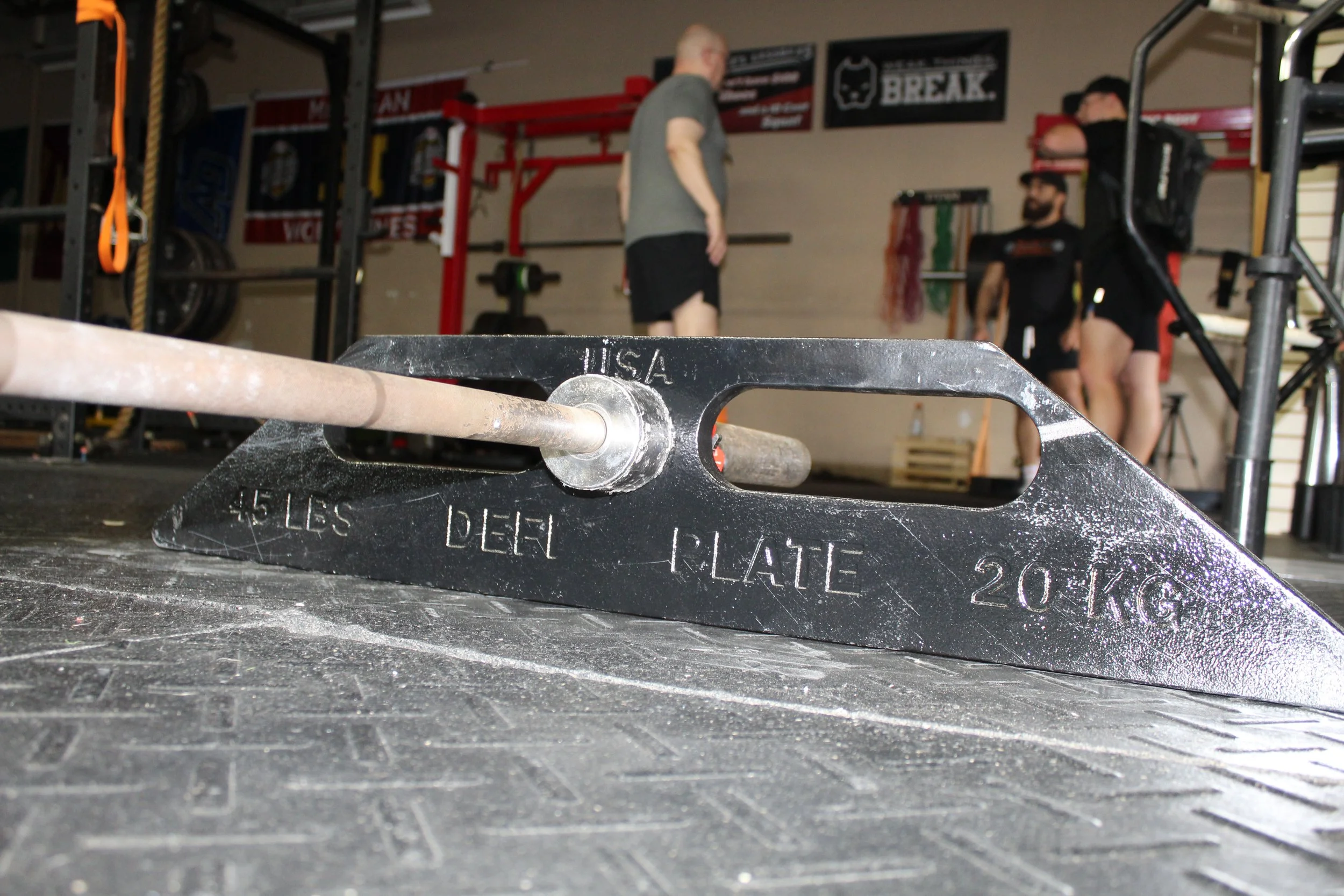 An american made USA weightlifting plate resting on a metal platform with weight plates, marked 45 lbs and 20 kg, in a gym with people in the background.