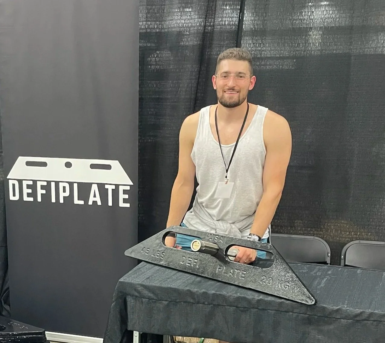 A man with a beard, glasses, and a sleeveless gray shirt standing behind a black table with a large black defibrillation paddles. There is a black curtain in the background and a sign with the word 'DEFIPLATE' and a graphic of versatile weight plate.