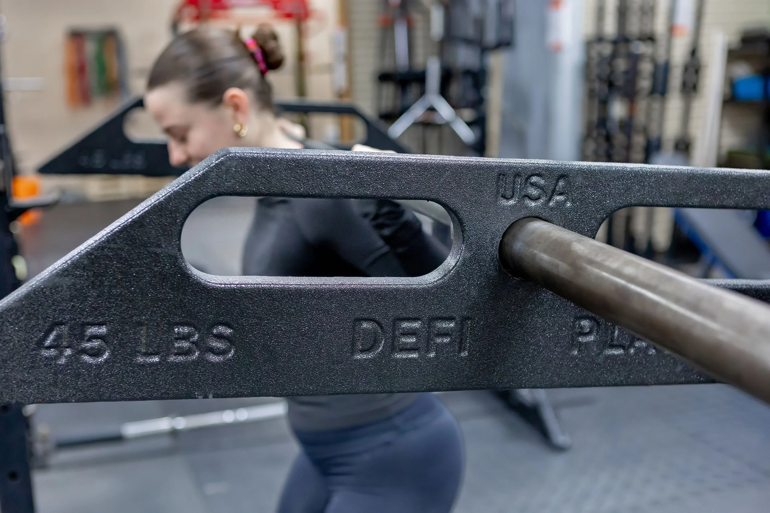 Close-up of a 45-pound black weight plate with a metal barbell in a gym, a woman in black workout clothes is blurred in the background.