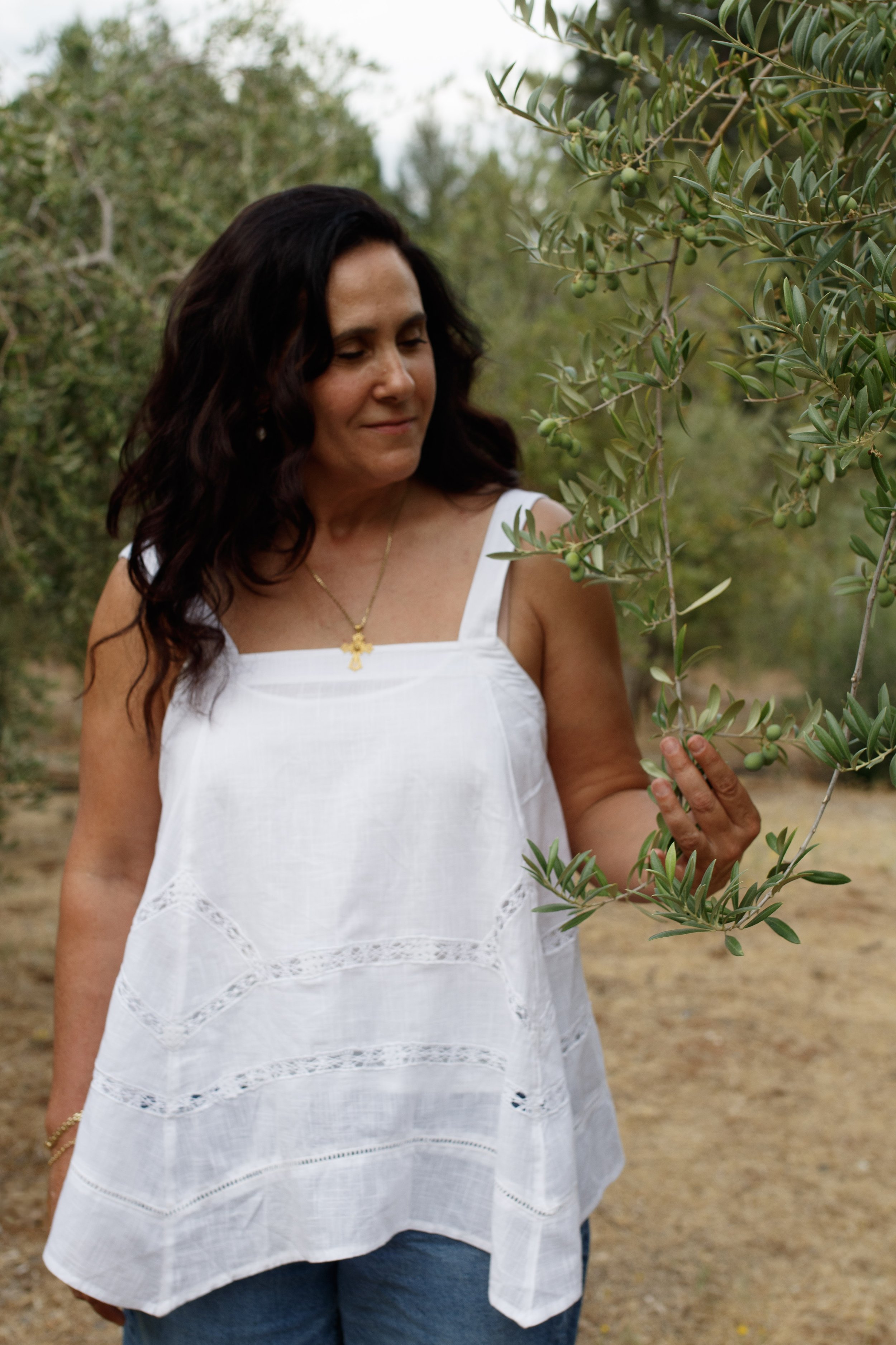 A woman with dark wavy hair wearing a white sleeveless summer dress with embroidered details, holding a branch of an olive tree, standing outdoors in a green orchard.