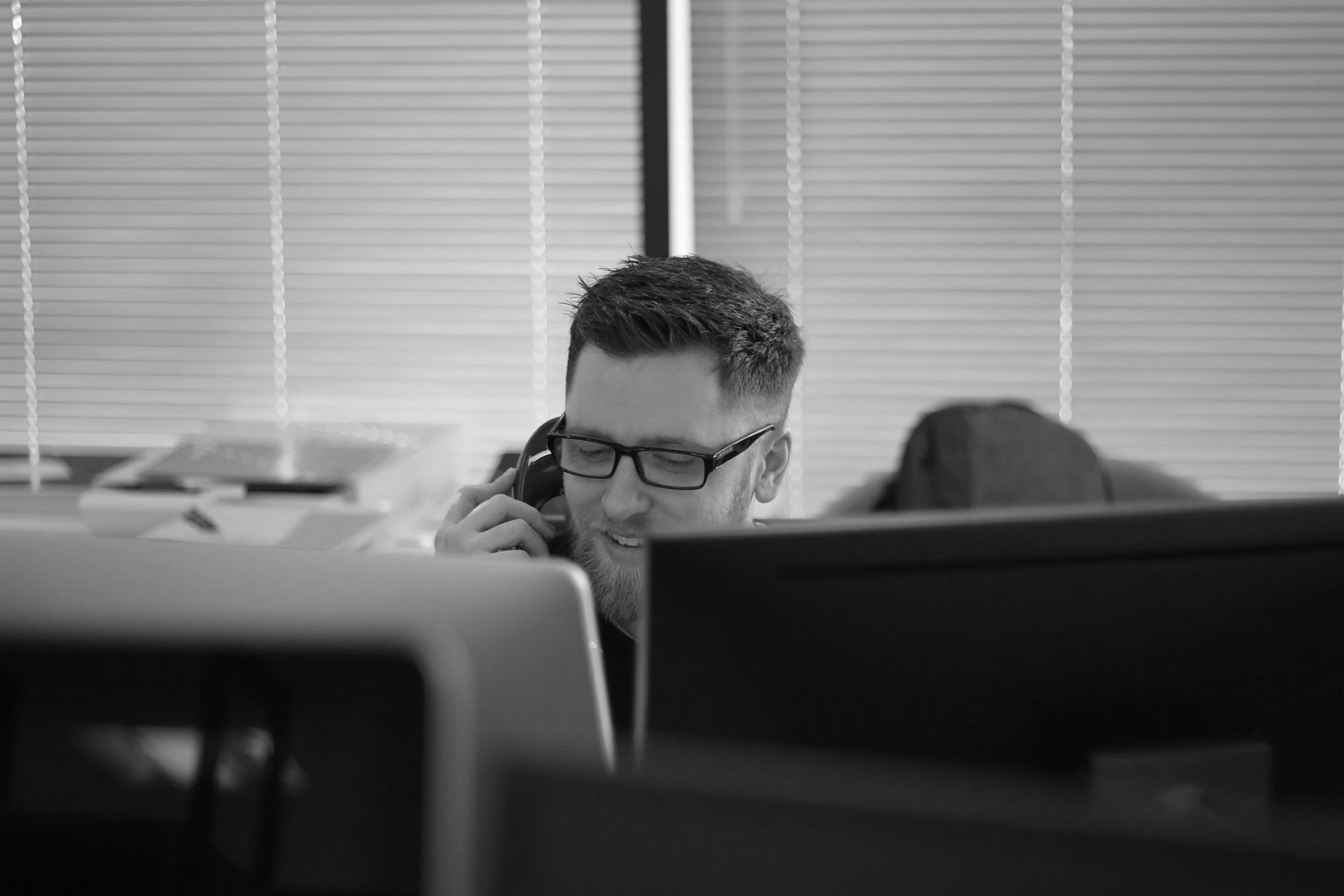 A man with glasses and a beard talking on a landline phone in an office, surrounded by computers and office supplies, with blinds covering the windows in the background.
