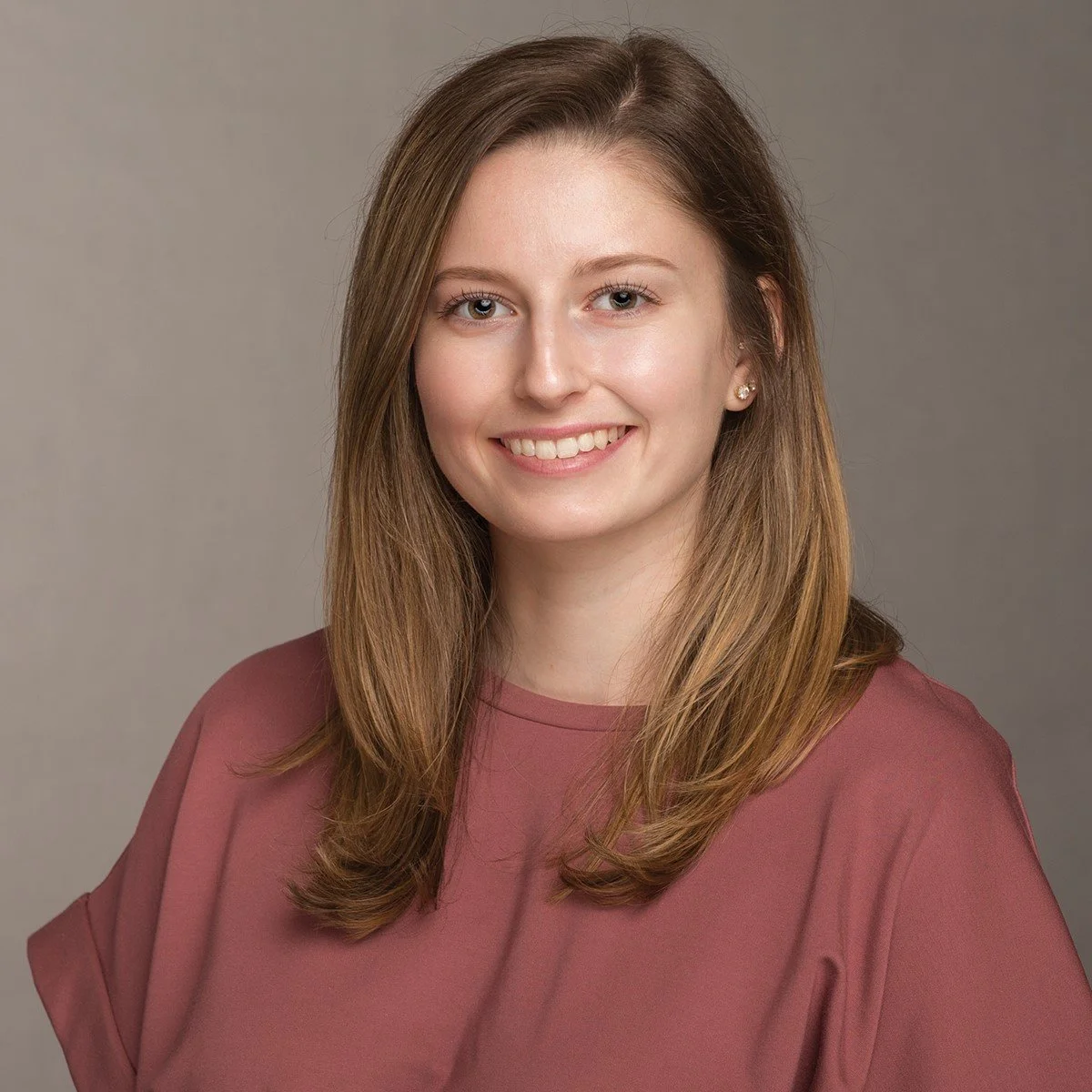 A young woman with shoulder-length light brown hair, blue eyes, and light skin, smiling at the camera against a neutral gray background, wearing a mauve shirt and small earrings.