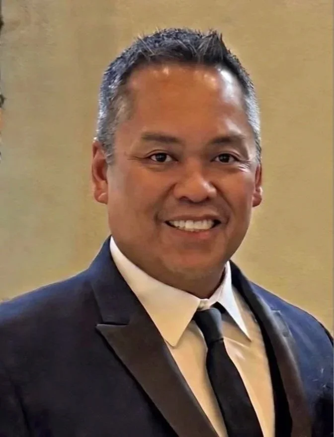 A smiling man with short, dark hair, dressed in a dark suit, white shirt, and black tie, standing in front of a beige background.