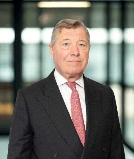Portrait of a man wearing a dark suit, white shirt, and red patterned tie, standing in front of a modern glass building in London.