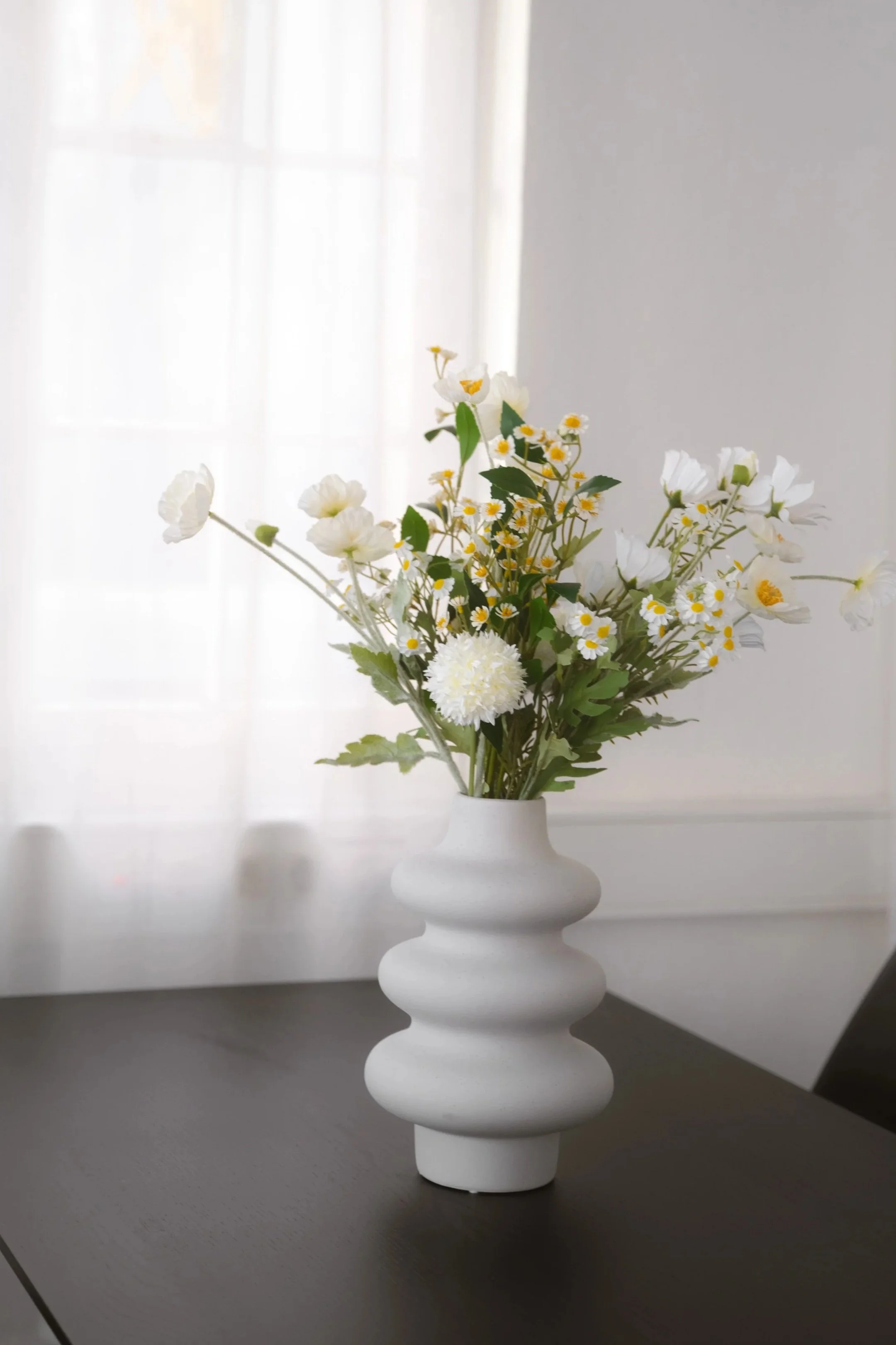Weiße Vase mit einer Blumenstrauß aus weißen und gelben Blumen, auf einem dunklen Tisch vor einem Fenster mit weißen Gardinen.