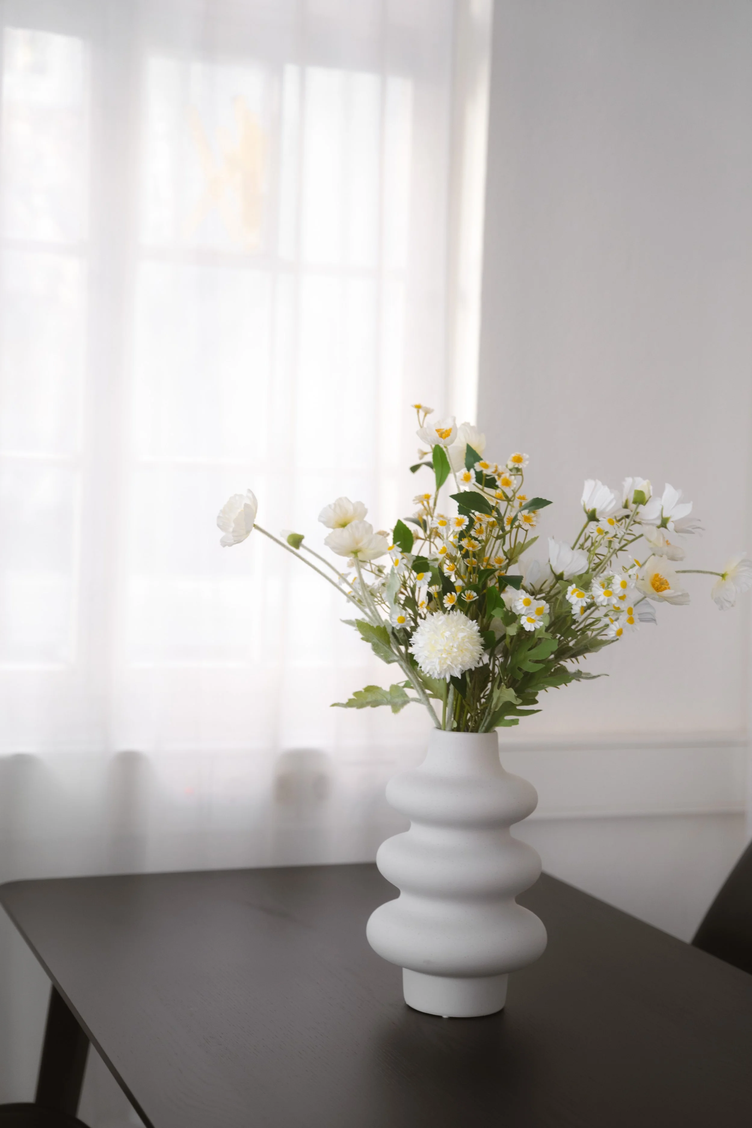 Weiße Keramikvase mit weißen und gelben Blumen auf einem dunklen Tisch vor einem Fenster mit halbtransparenten Vorhängen.