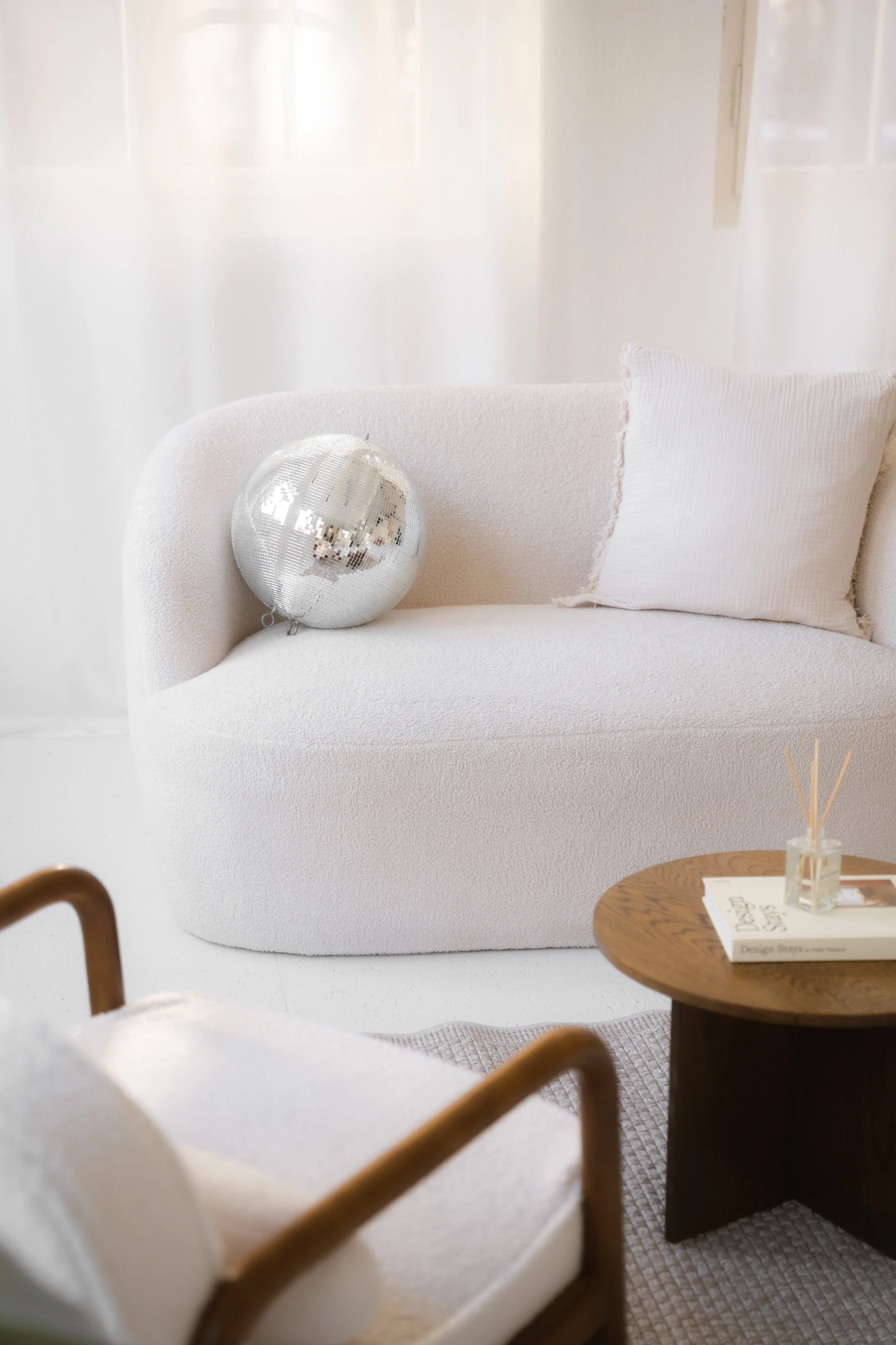 Moderner, weißer Couch mit Kissen, ein silberner Disco-Ball auf der Armlehne, ein runder Holztisch mit Bücher und einem Diffusor, im Hintergrund weiße Vorhänge.