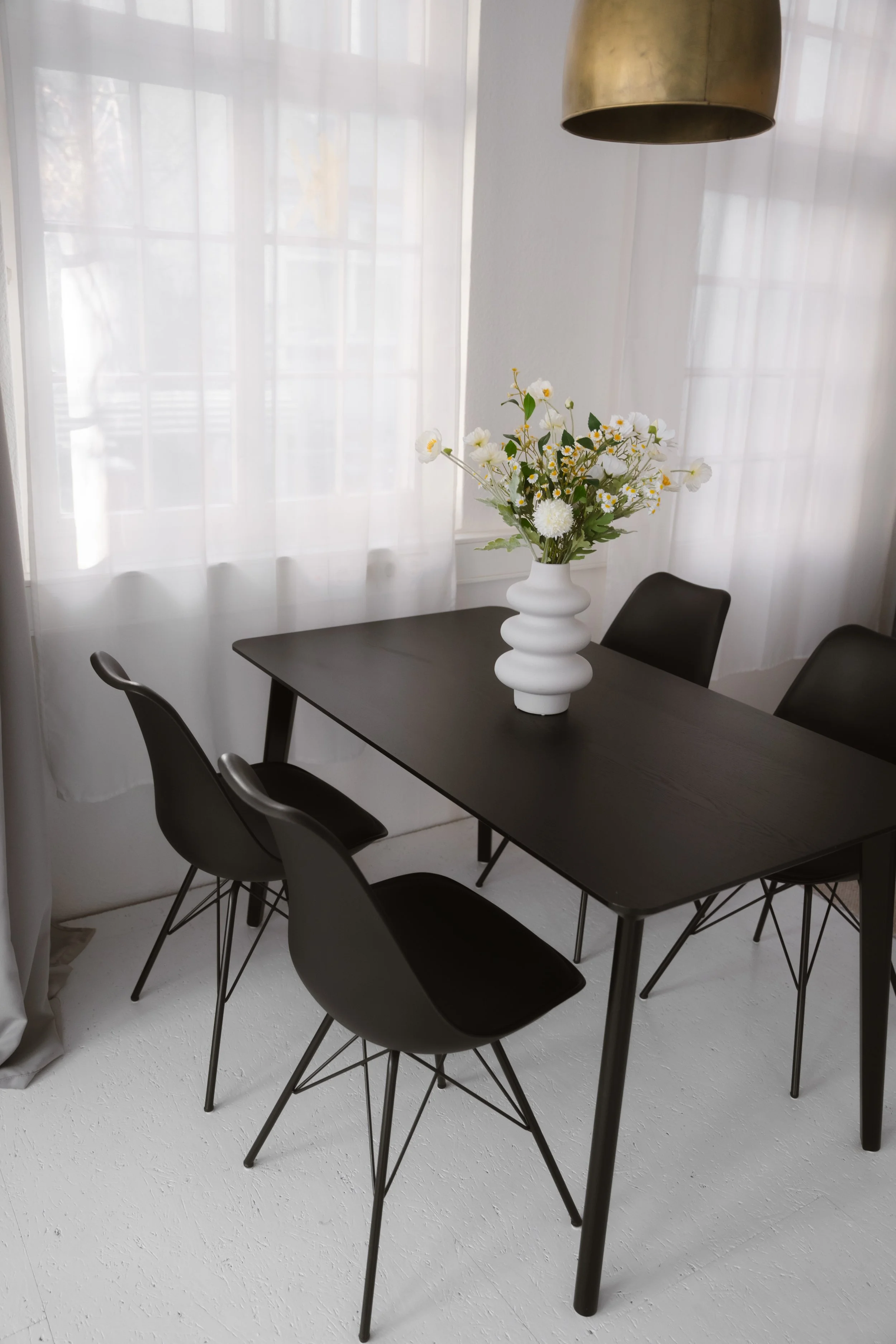 Ein schwarzer Esstisch mit vier schwarzen Stühlen, eine weiße Vase mit weißen Blumen auf dem Tisch, Fenster mit weißen Vorhängen im Hintergrund, eine große goldene Hängeleuchte oben.