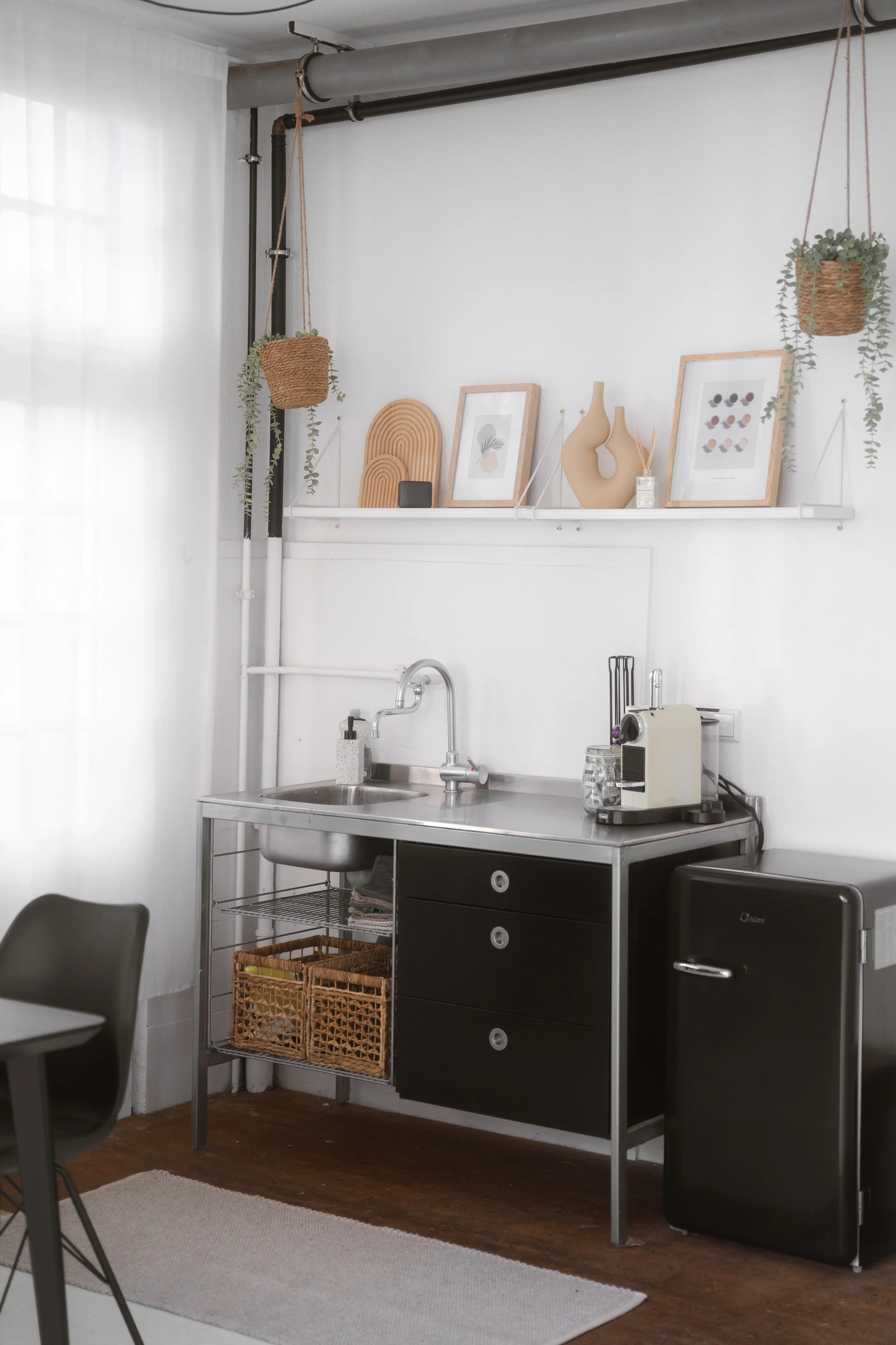 Küchenbereich mit dunklem Unterschrank, Spüle, Wasserhahn, Kaffeemaschine, Wäschekorb und Wandregal mit Dekorationsgegenständen und Bildern, natürlicher Licht durch Fenster, teurer Holzboden.