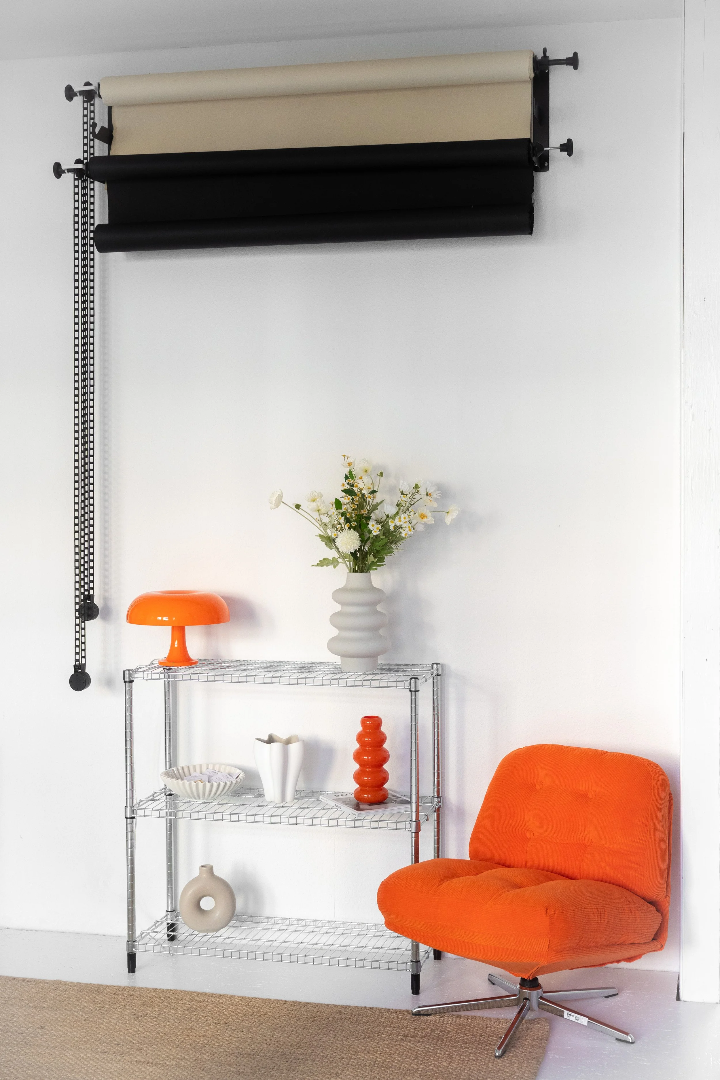 Ein Raum mit einem weißen Wandhintergrund, einer Metallregal mit Dekorationsvasen und einer leuchtend orangefarbenen Sitzgelegenheit, darüber eine Wanddekoration mit Rollstoffen in Beige und Schwarz.