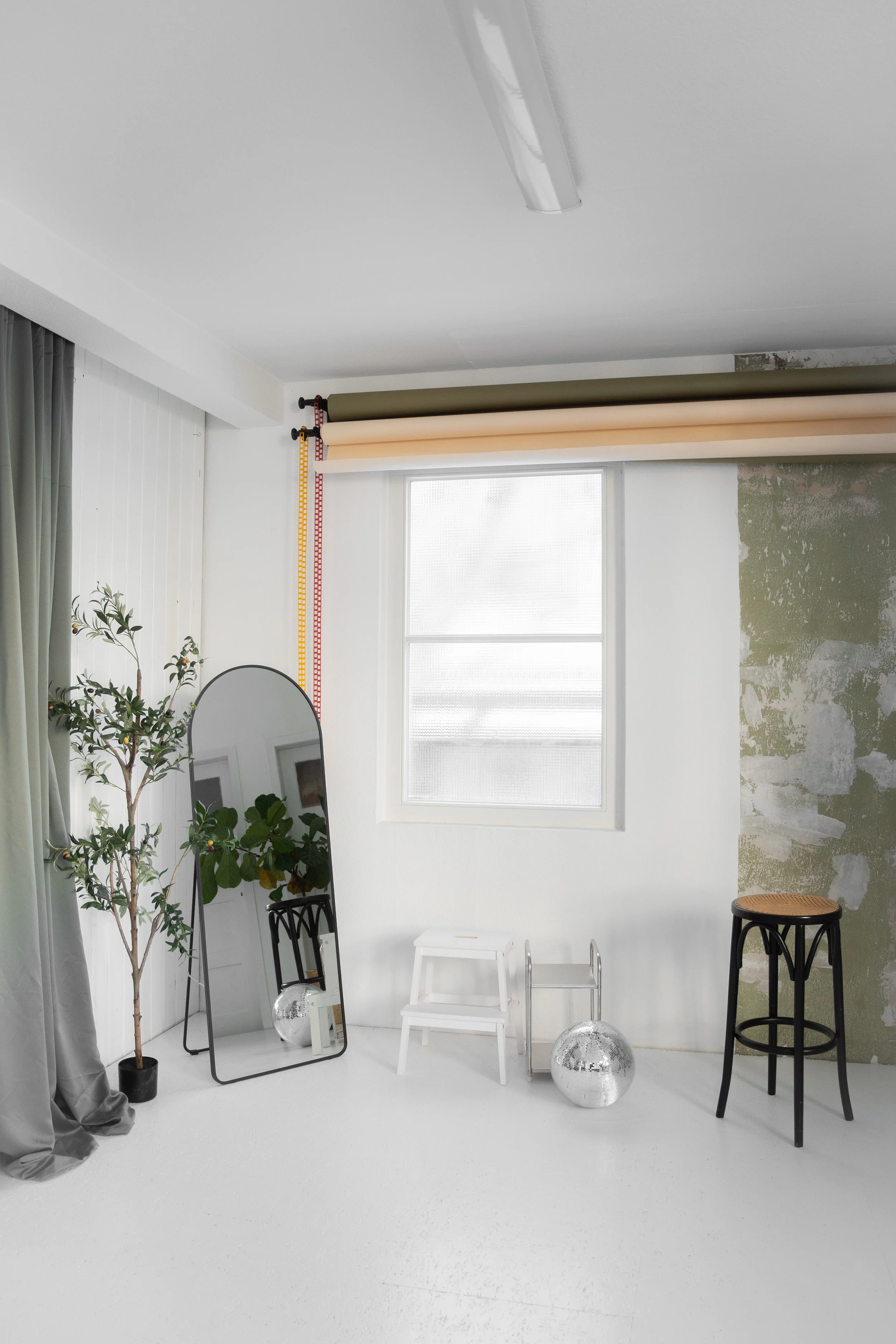 Ein weißer Raum mit einem Fenster, einer Stehlampe, einem Wandspiegel, einer Pflanze, einem kleinen weißen Stuhl, einem Hocker und zwei Disco-Kugeln.