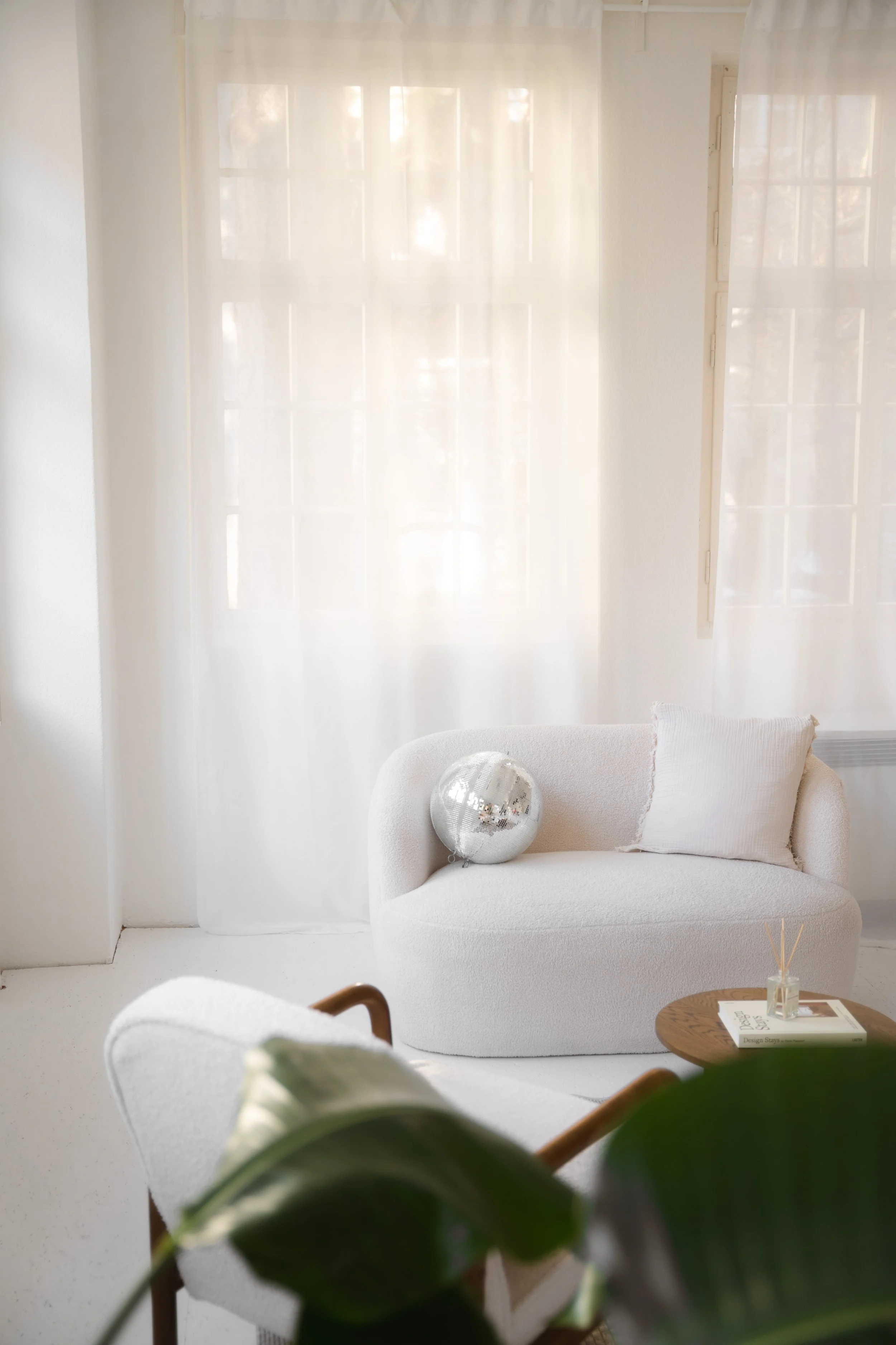 Ein heller, minimalistisch eingerichteter Raum mit weißen Vorhängen, einer weißen Couch mit Kissen, einem kleinen runden Holz-Tisch mit Büchern und einem Duftstäbchen-Glas. Ein silberner Ballon liegt auf der Couch, im Vordergrund ist eine grüner Pfla