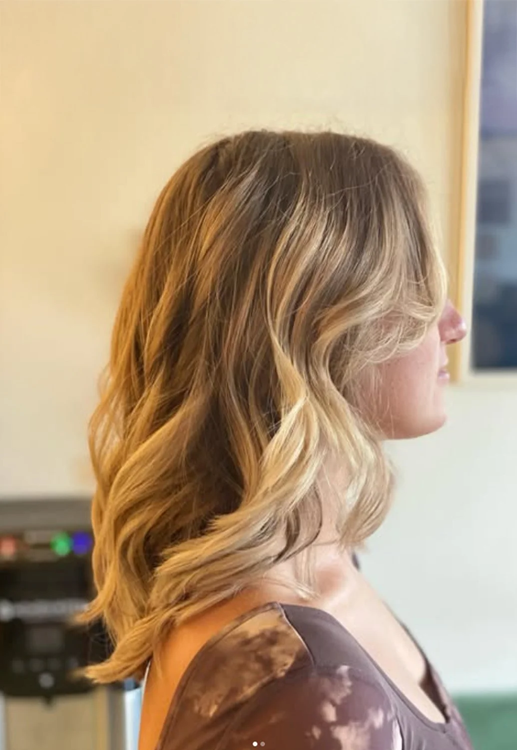 Side profile of a woman with shoulder-length wavy blonde hair, wearing a brown patterned top, standing indoors near a window.