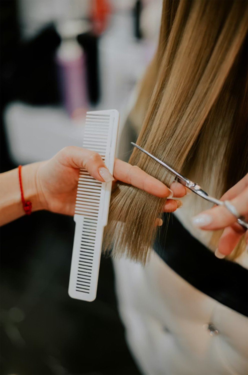 A hairdresser is cutting a woman's long blonde hair with scissors while holding a white comb and hair clipper.