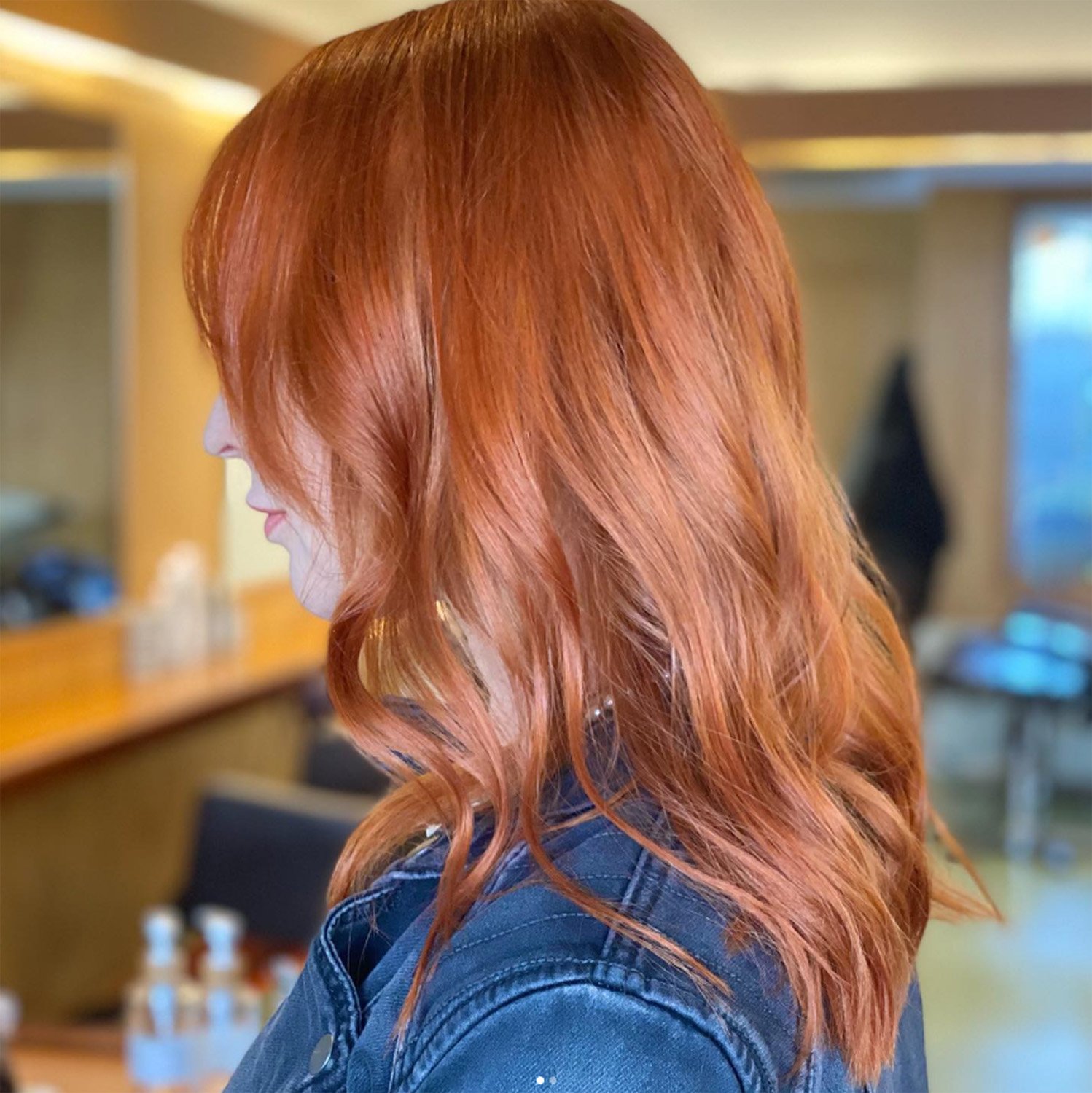 Close-up of a woman with long, wavy red hair, wearing a denim jacket, inside a room with wooden and blue furniture and a mirror in the background.