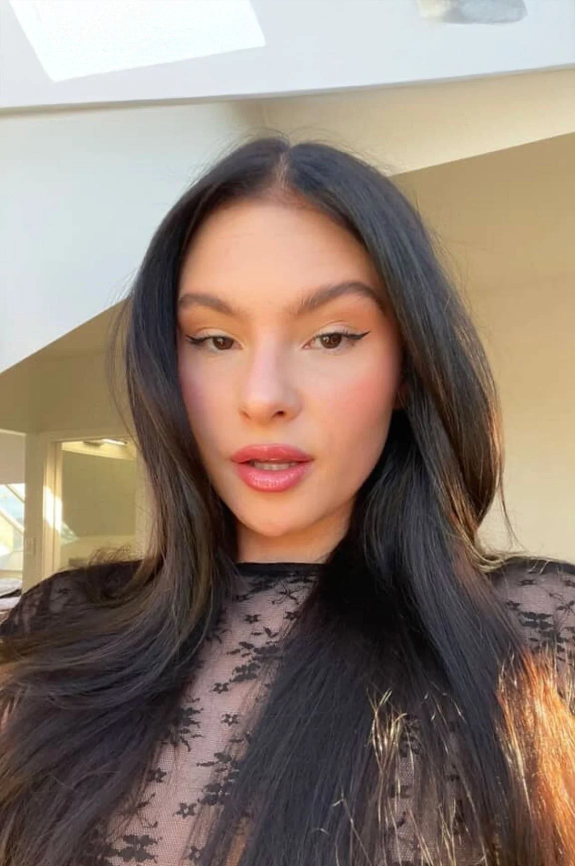 A woman with long black hair, wearing makeup, and a black lace top, standing indoors near a window with natural light.