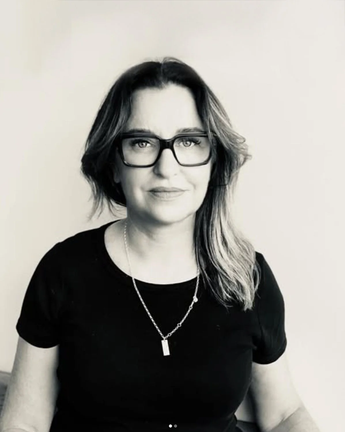 Black and white photo of a woman with shoulder-length wavy hair, wearing glasses, a black top, and a necklace, sitting against a plain light-colored background.