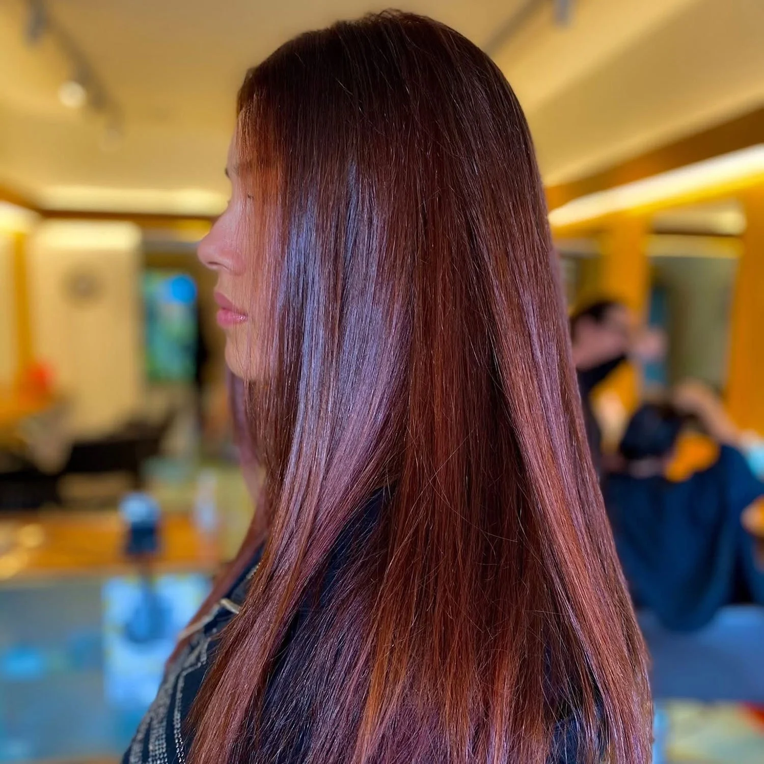 Side view of a woman with long, straight, reddish-brown hair, inside a brightly lit salon with yellow and blue decor.