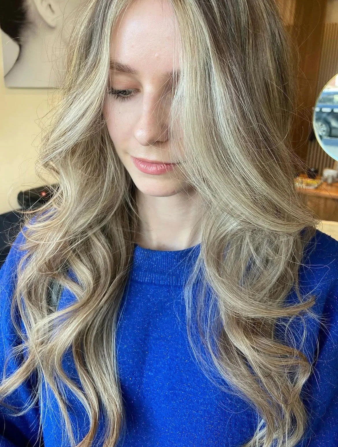 A young woman with long, wavy blonde hair wearing a blue top, looking downward and slightly smiling in an indoor setting.