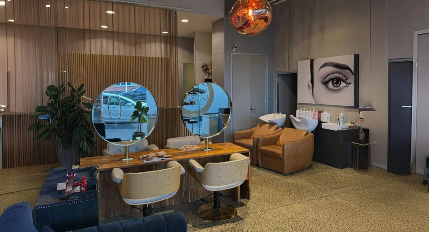 A modern salon with a wooden partition, two mirrors on a wooden table, and seating with chairs and armchairs. There are hair washing basins and a large black-and-white eye portrait on the wall.
