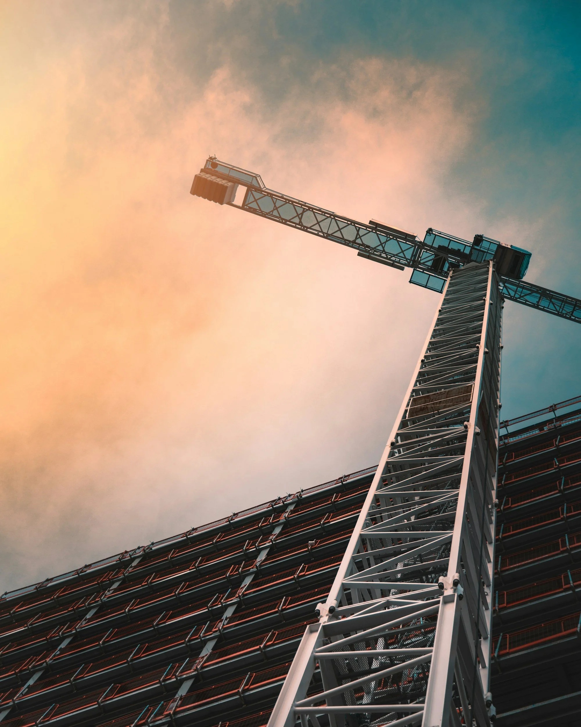 A construction crane tower extending into a sky with orange and blue hues.