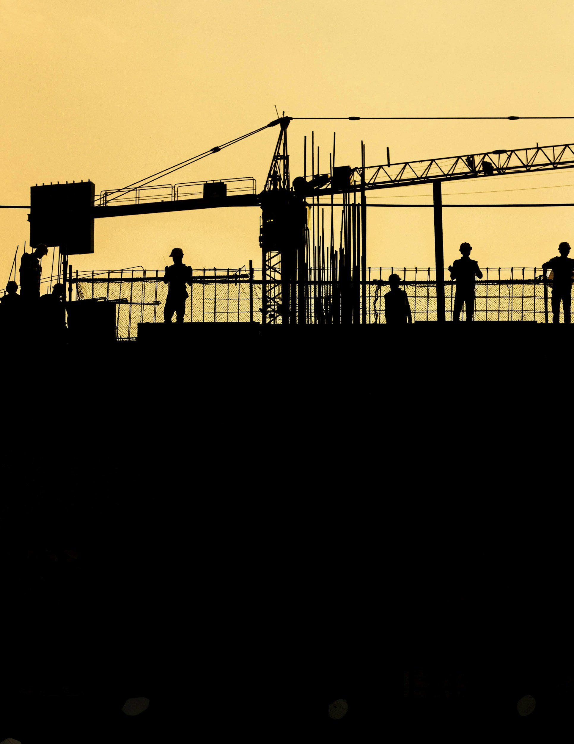 Silhouettes of construction workers on a building site with a crane against a yellow sky at sunset or sunrise.
