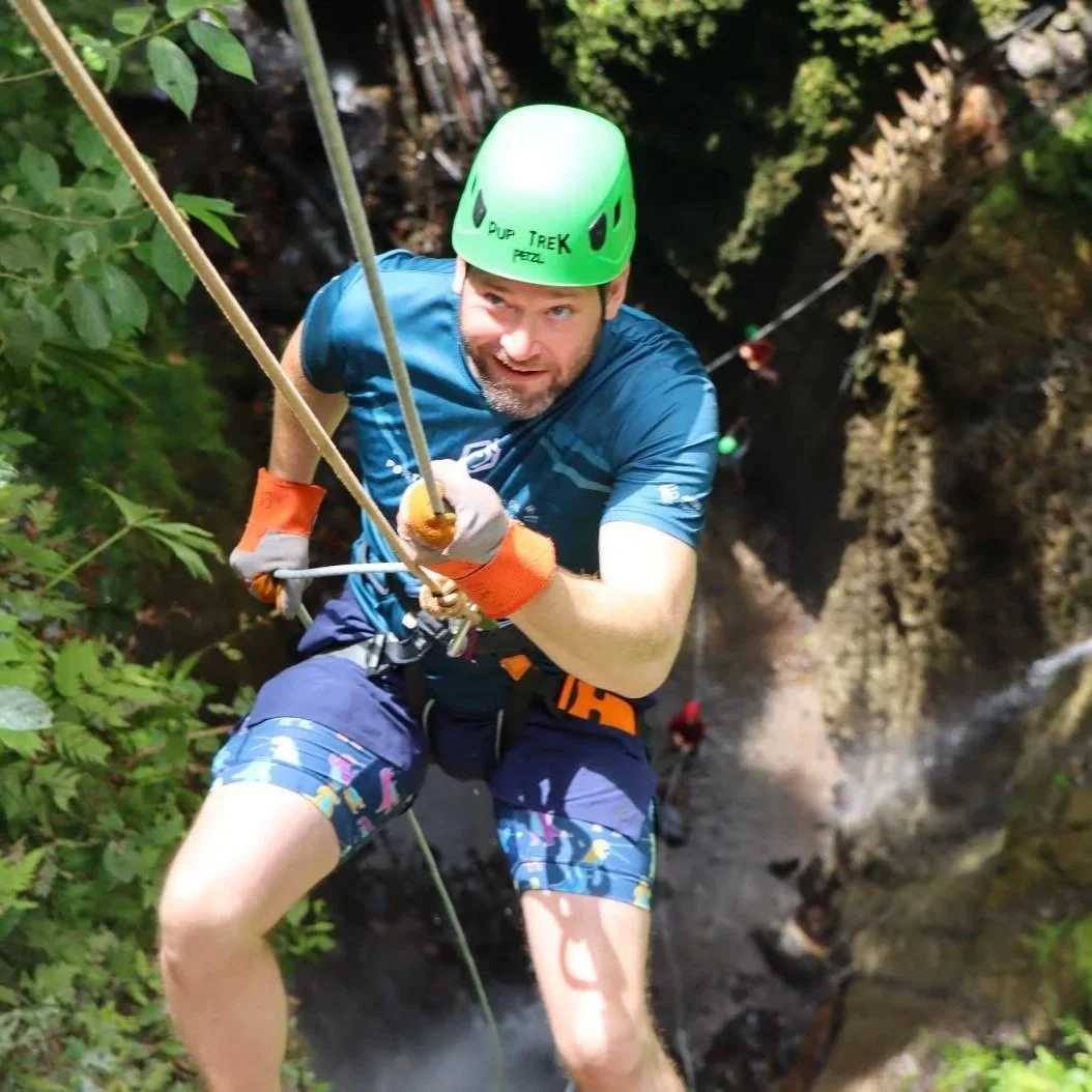 A man wearing a green helmet and blue shirt climbing a rock wall while holding onto ropes, surrounded by green foliage.