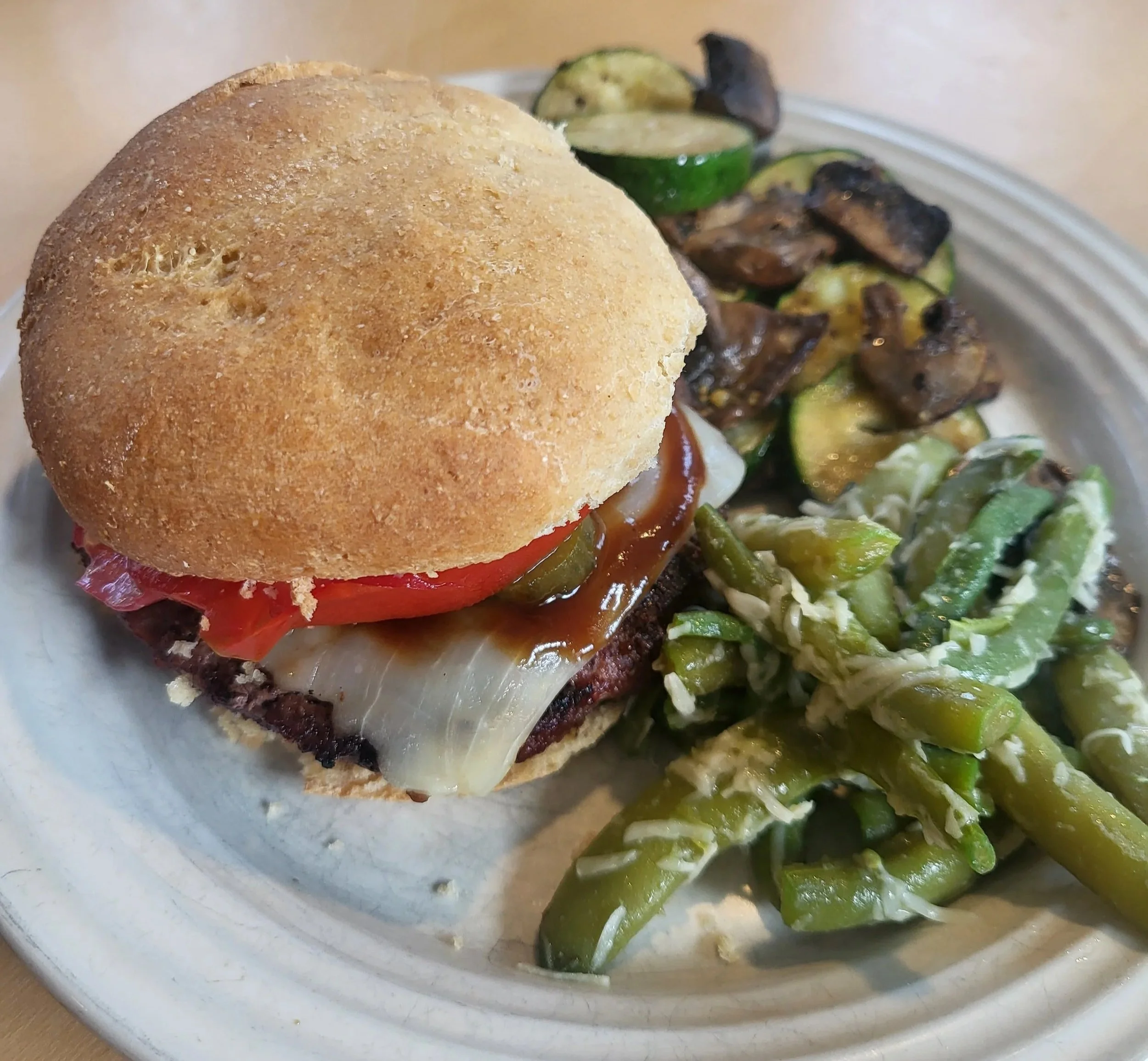 Close-up of a hamburger on a whole grain sandwich bun on a plate with grilled vegetables including zucchini and green beans on the side.