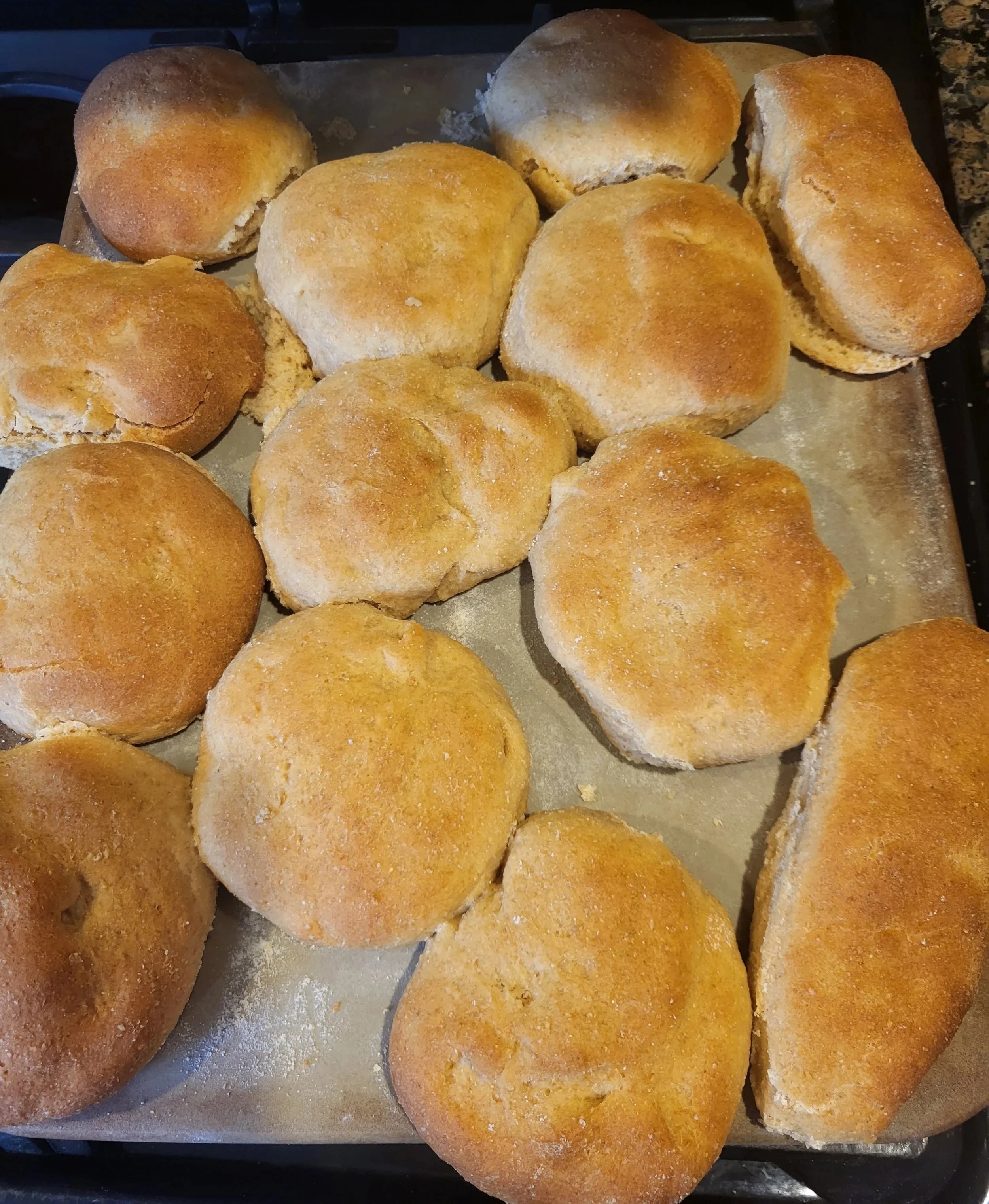 Freshly baked round bread rolls on a baking sheet, some with a light dusting of flour.