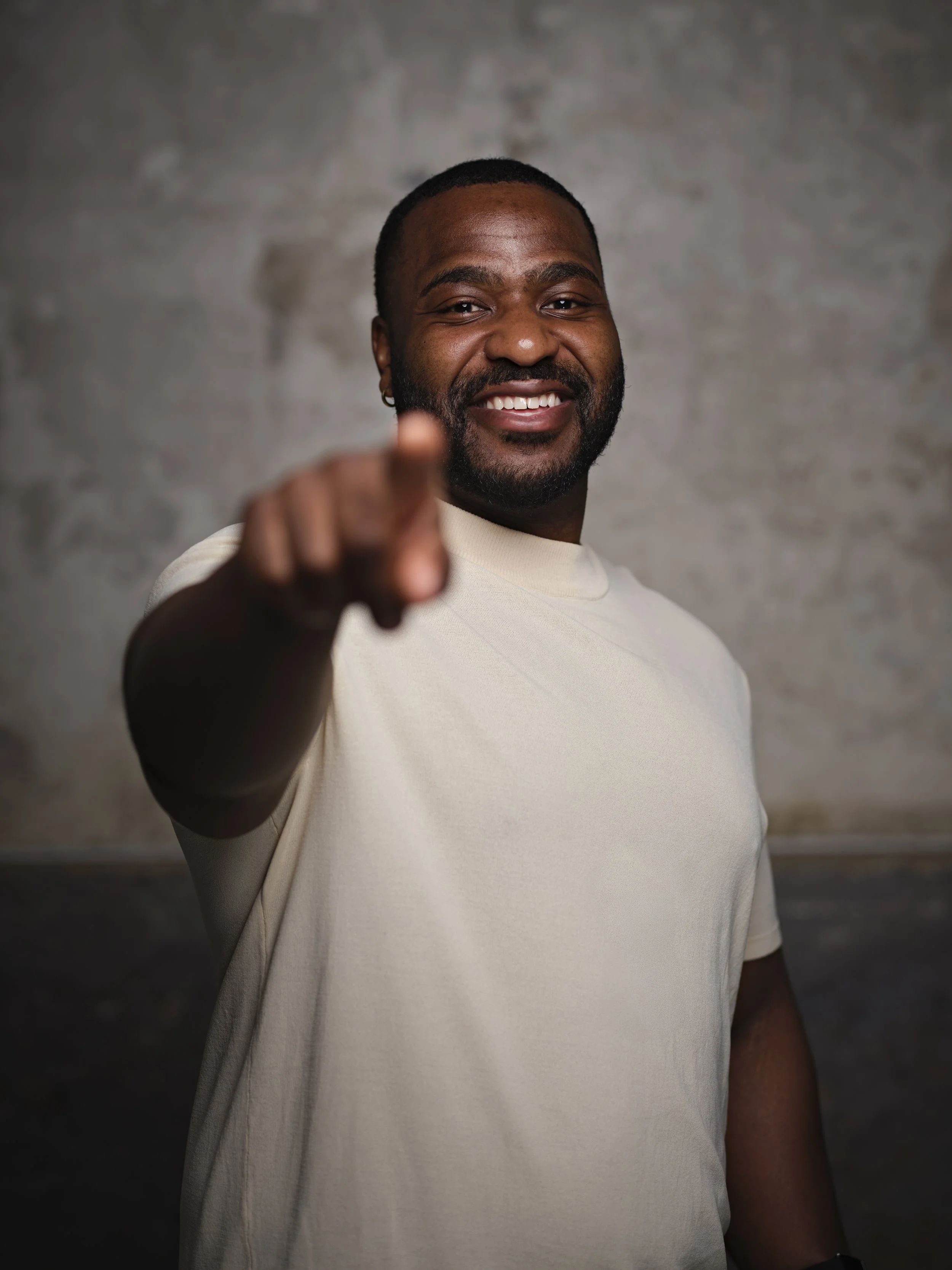 A smiling man with a beard and short hair pointing at the camera, wearing a light-colored t-shirt, standing against a plain background.
