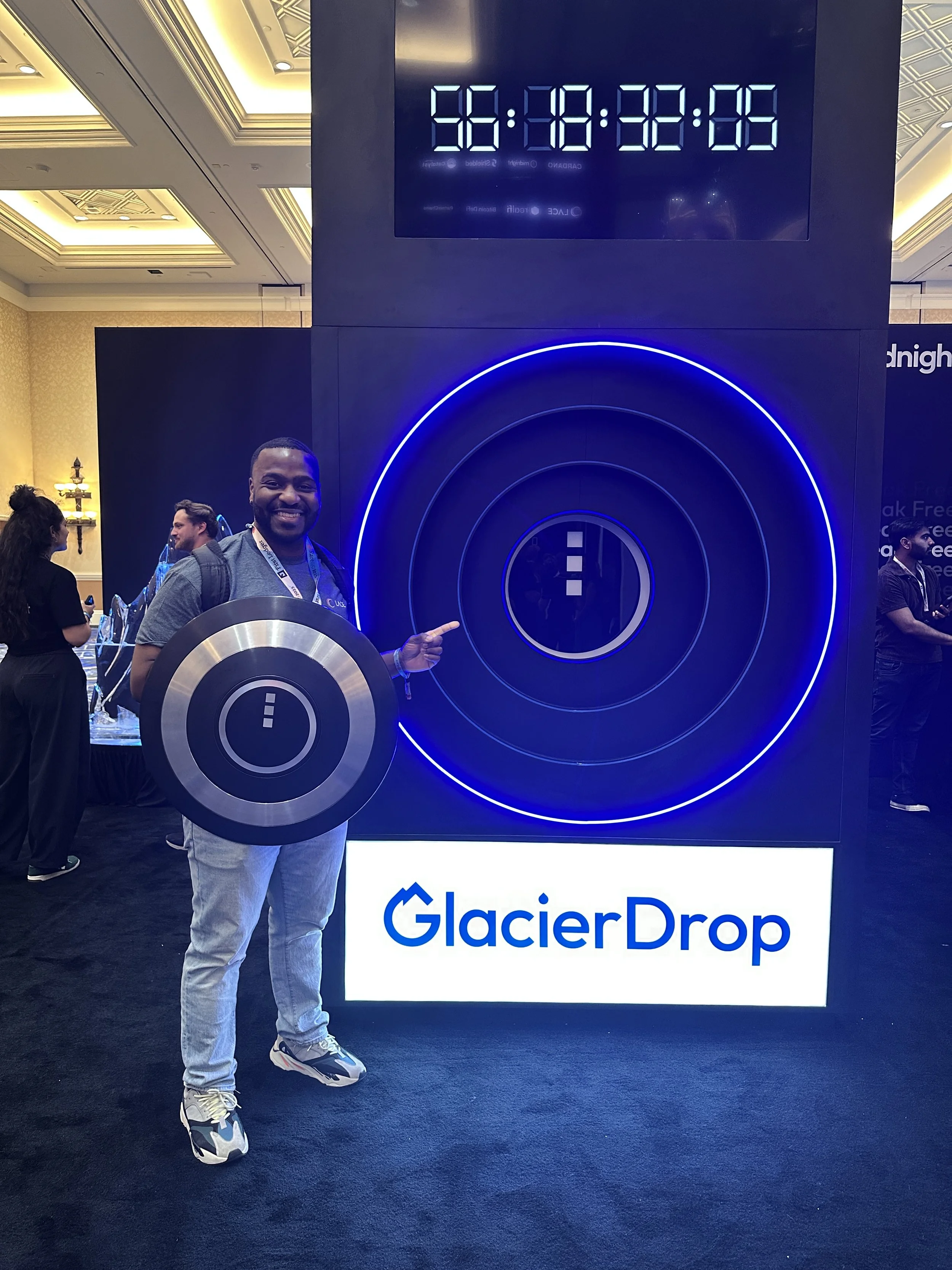 A man smiling and pointing at a large, illuminated circular graphic at a GlacierDrop event, holding a circular sign with a plus and minus symbol. The display shows a countdown timer.